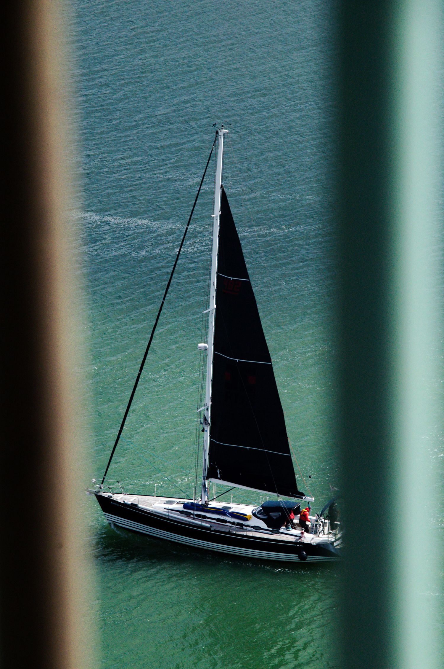 A sailboat with black sails glides through turquoise waters, viewed through a narrow gap in a green structure.