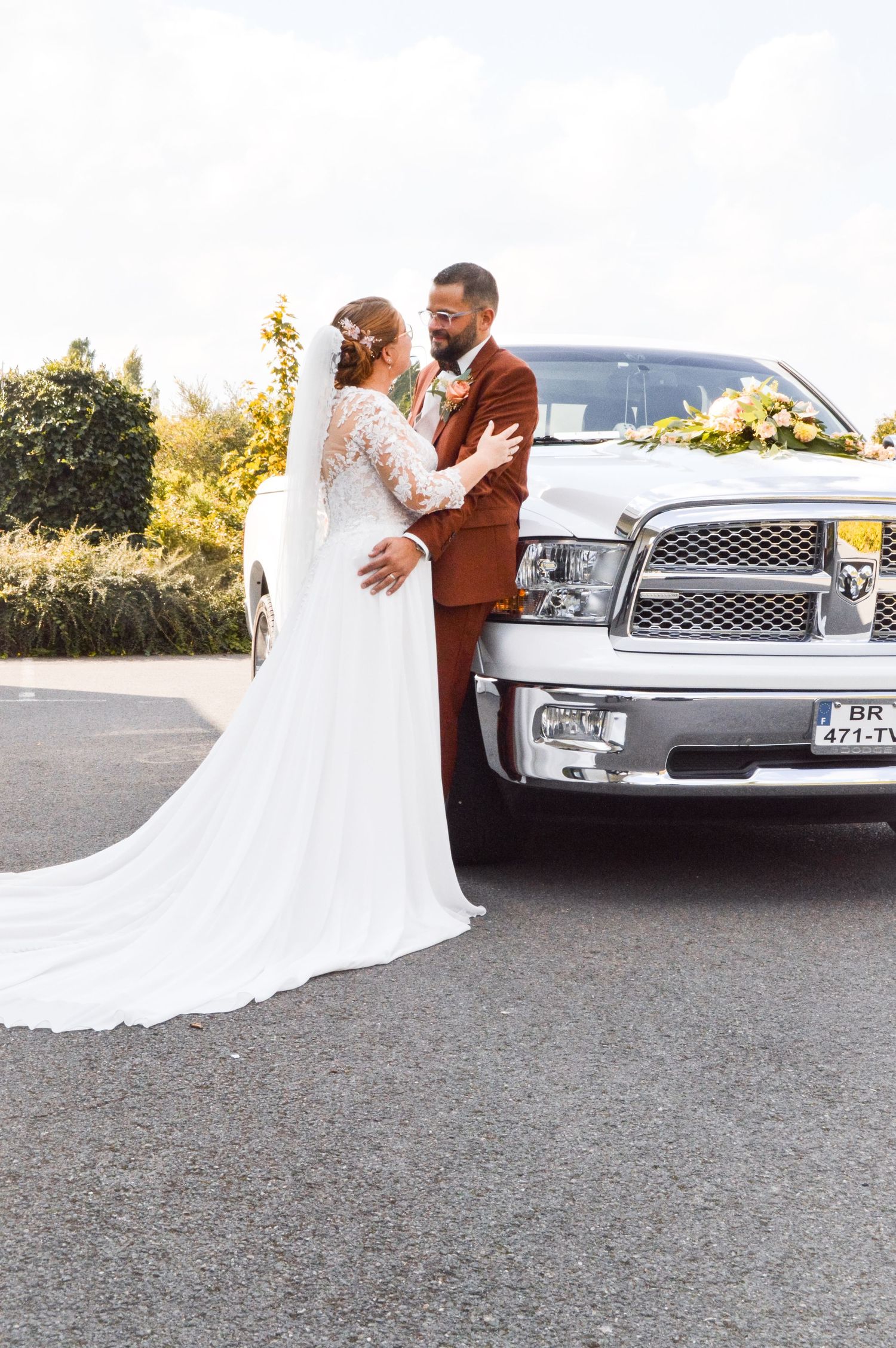 Couple in wedding attire posing by white limousine on sunny day.