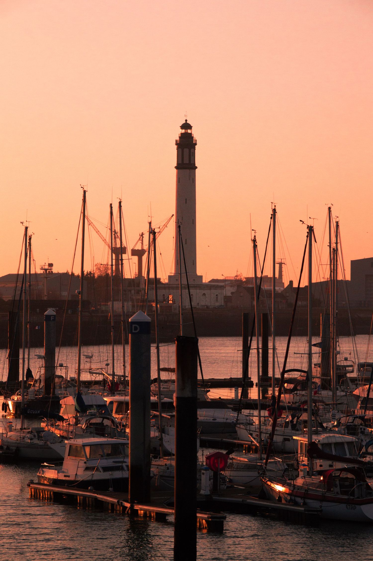 Sailboats docked at a marina during sunset with lighthouse silhouette and orange sky in the background.
