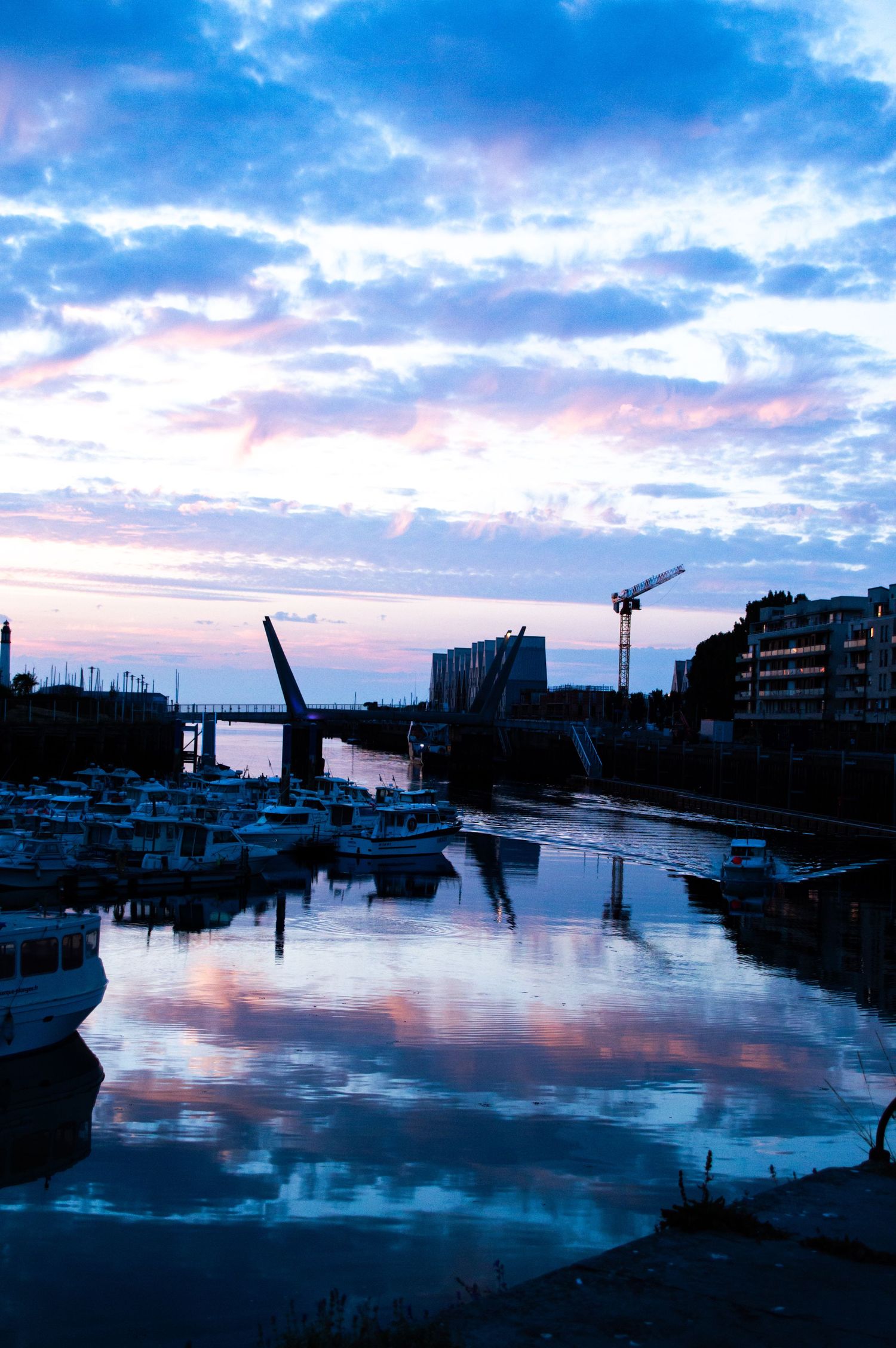 Twilight harbor with boats docked along calm reflective water, construction crane, drawbridge silhouette, and colorful pink-blue cloudy sky.