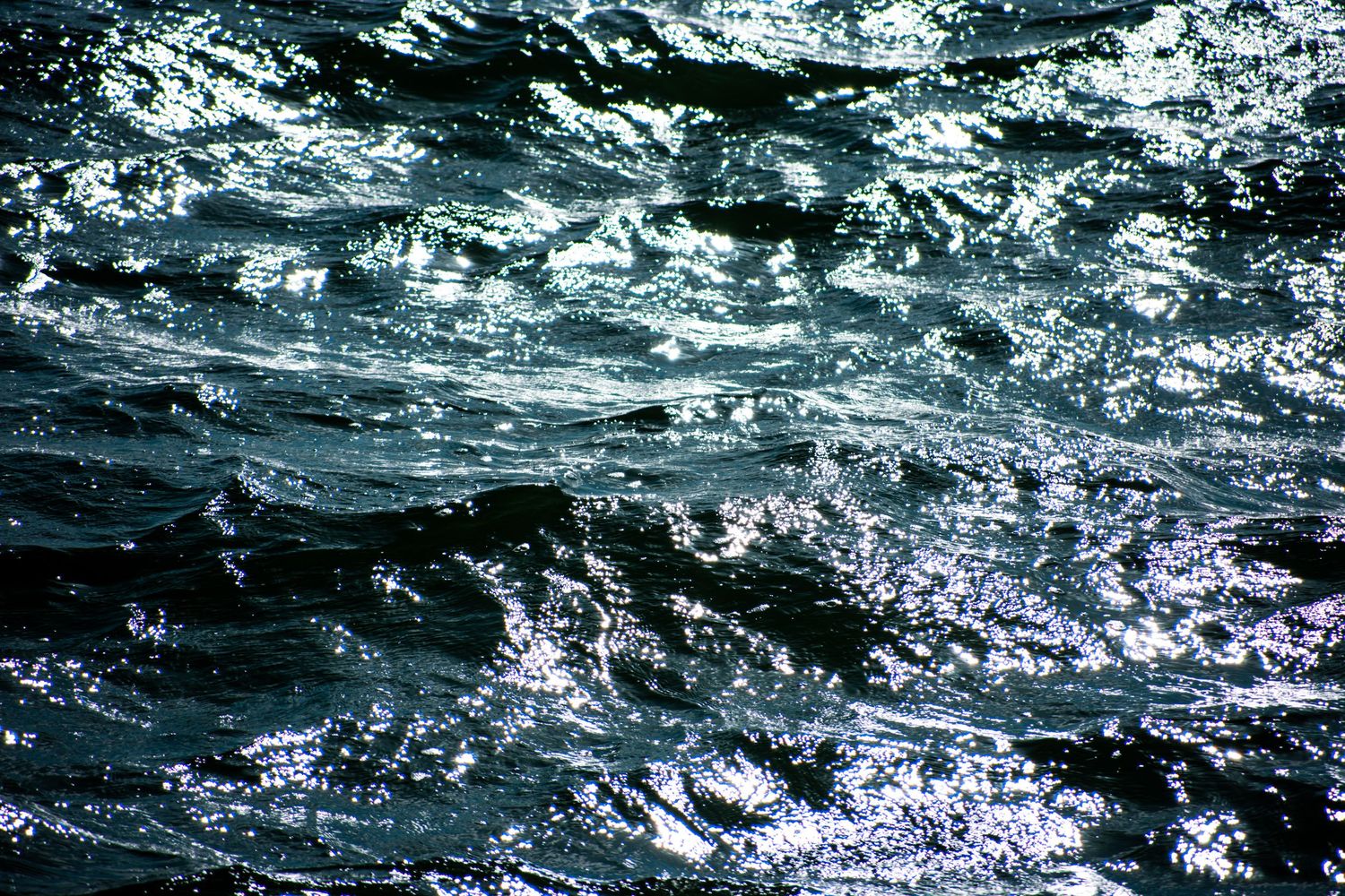Dark blue ocean waves with sunlight reflections creating a dramatic, turbulent water surface texture.
