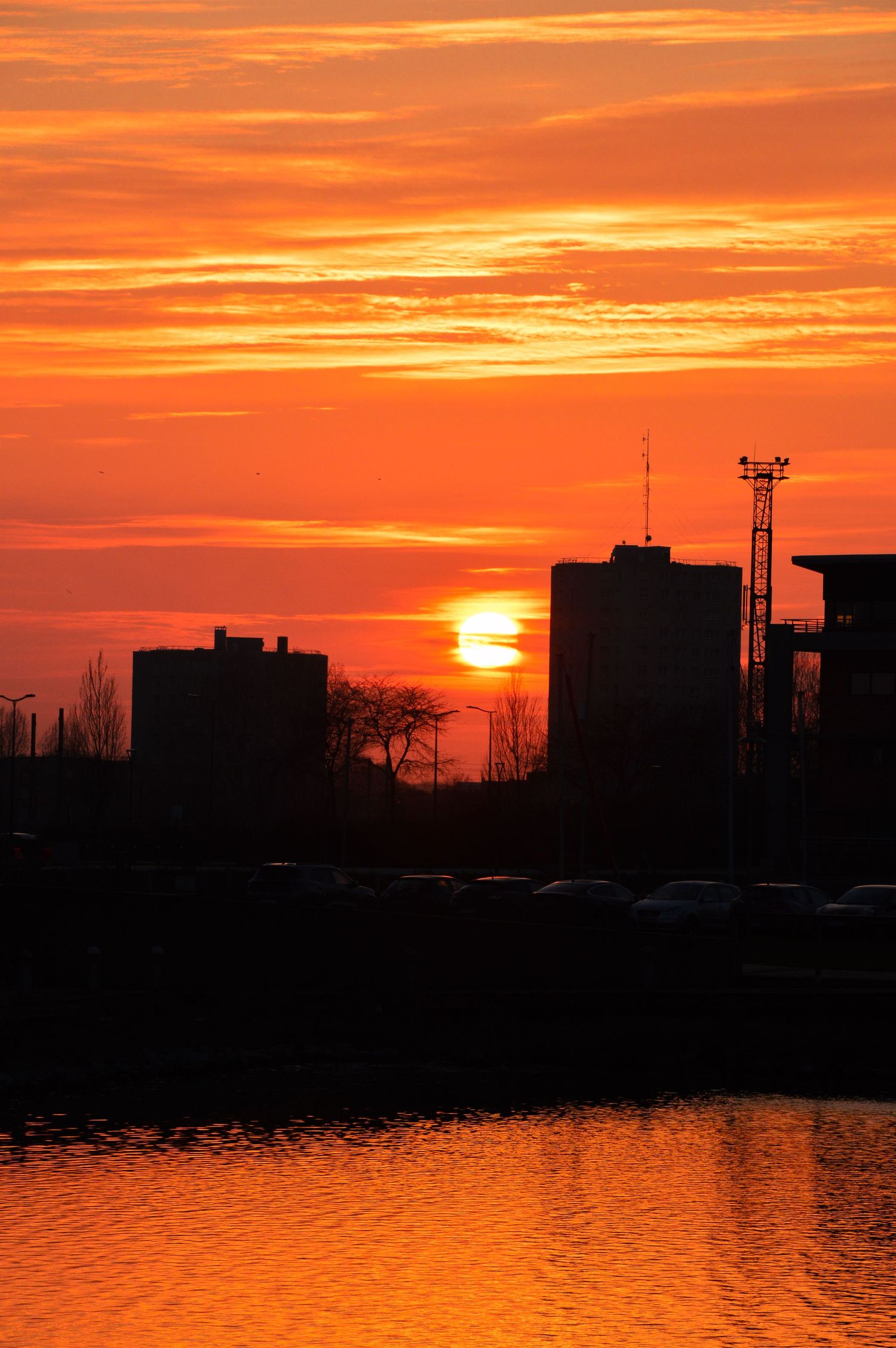 Ligne d’horizon urbaine avec des bâtiments et des antennes en silhouette sur un coucher de soleil orange au-dessus de l’eau.