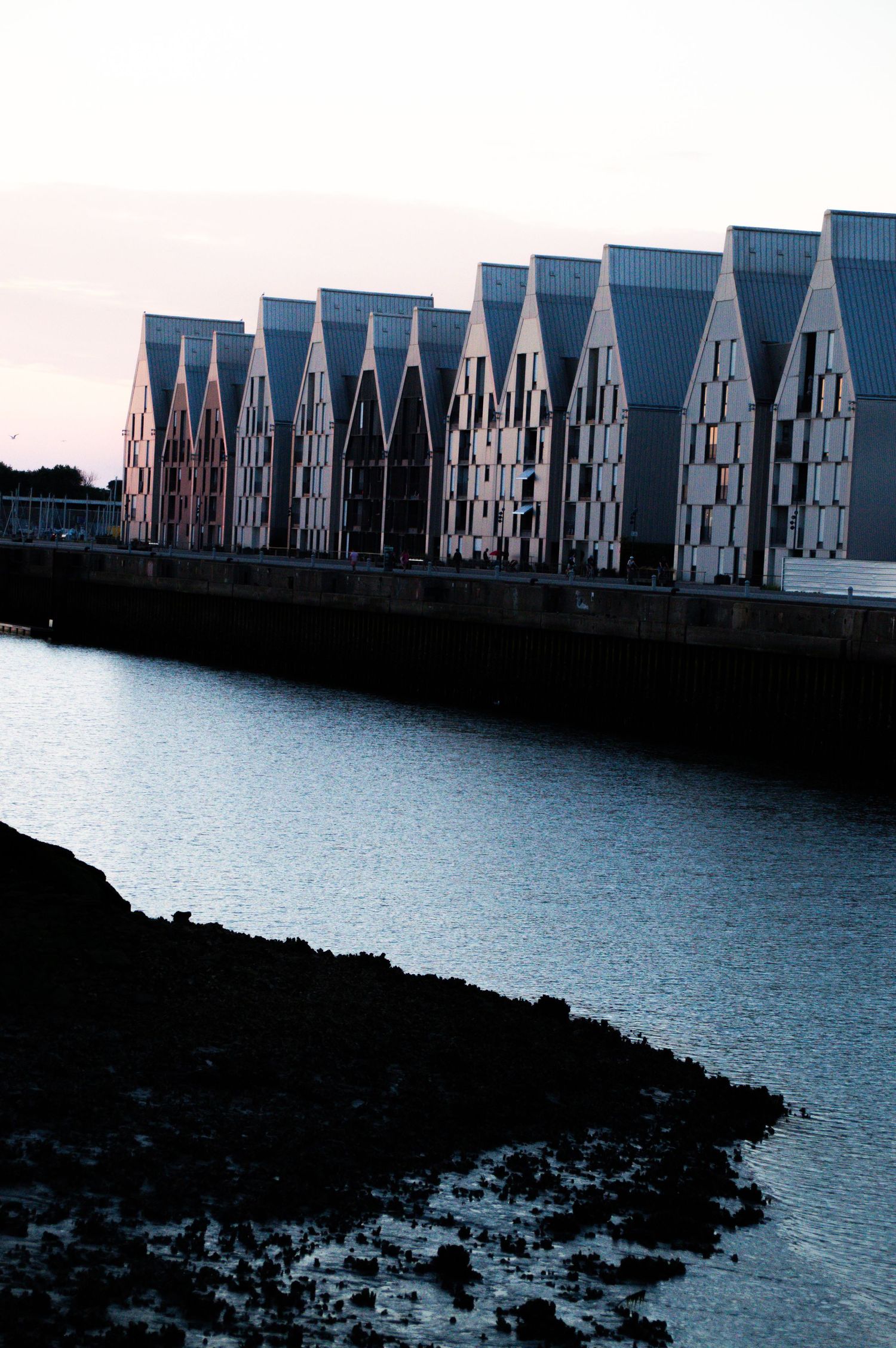 Modern waterfront buildings with pointed rooftops reflect on calm water at dusk in a coastal architectural complex.