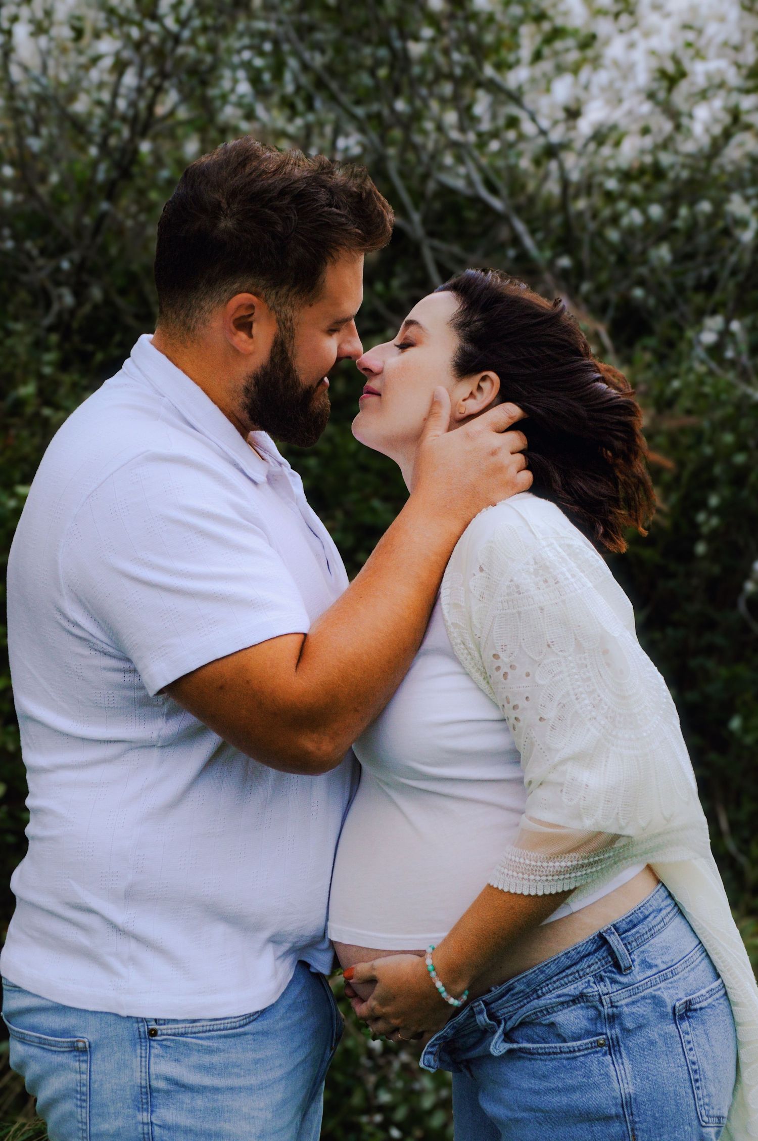 Intimate couple in casual attire embracing against flowering bushes.