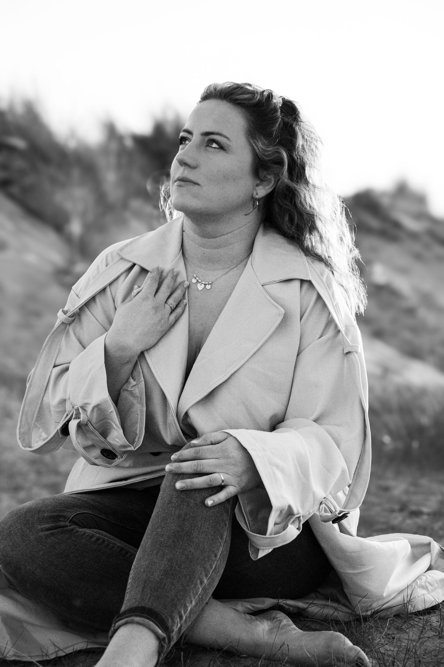 Black and white portrait of a woman sitting outdoors, hand on chest, gazing upward thoughtfully.
