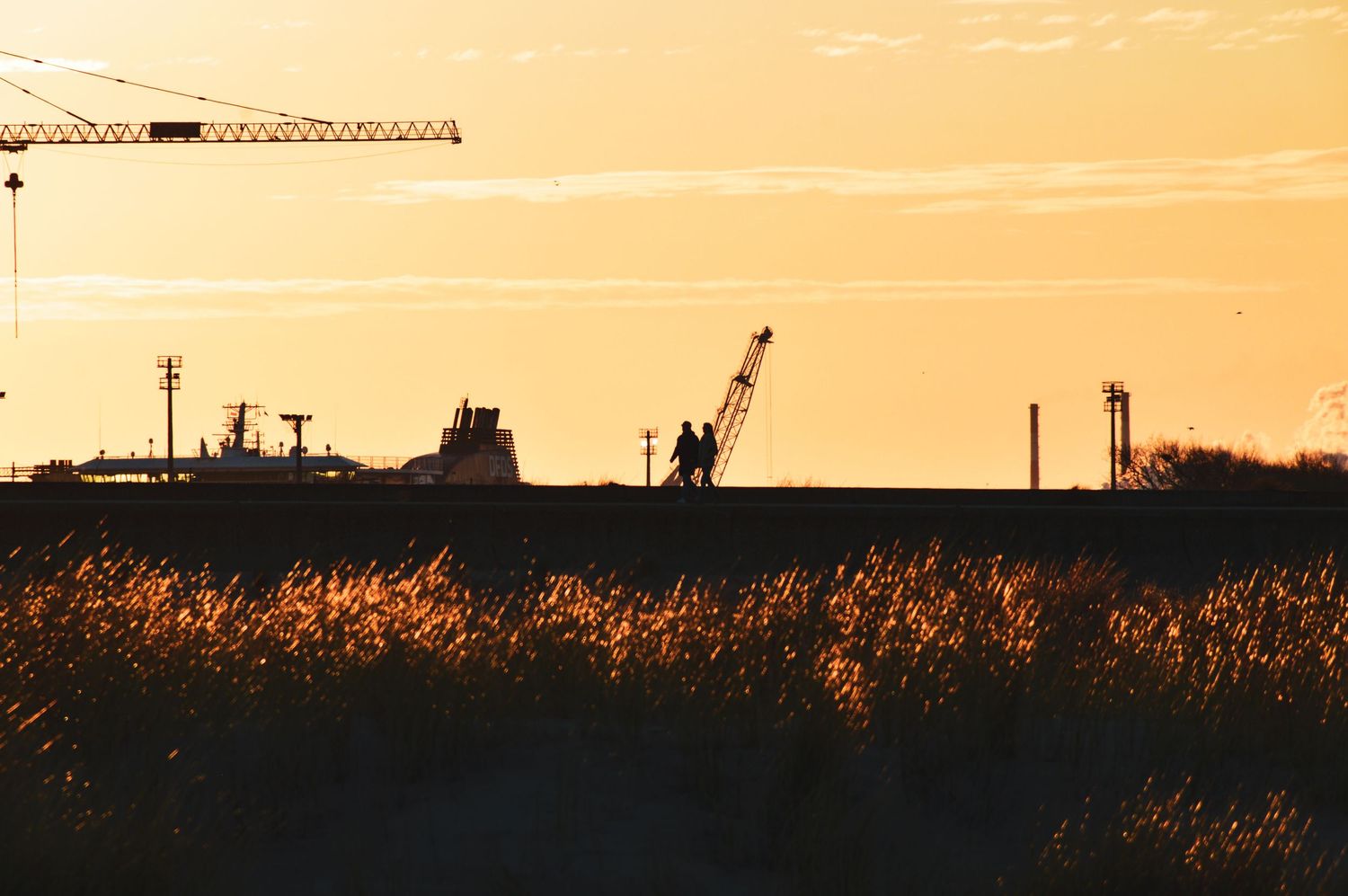 Industrial harbor silhouetted against an orange sunset sky with cranes and equipment visible.