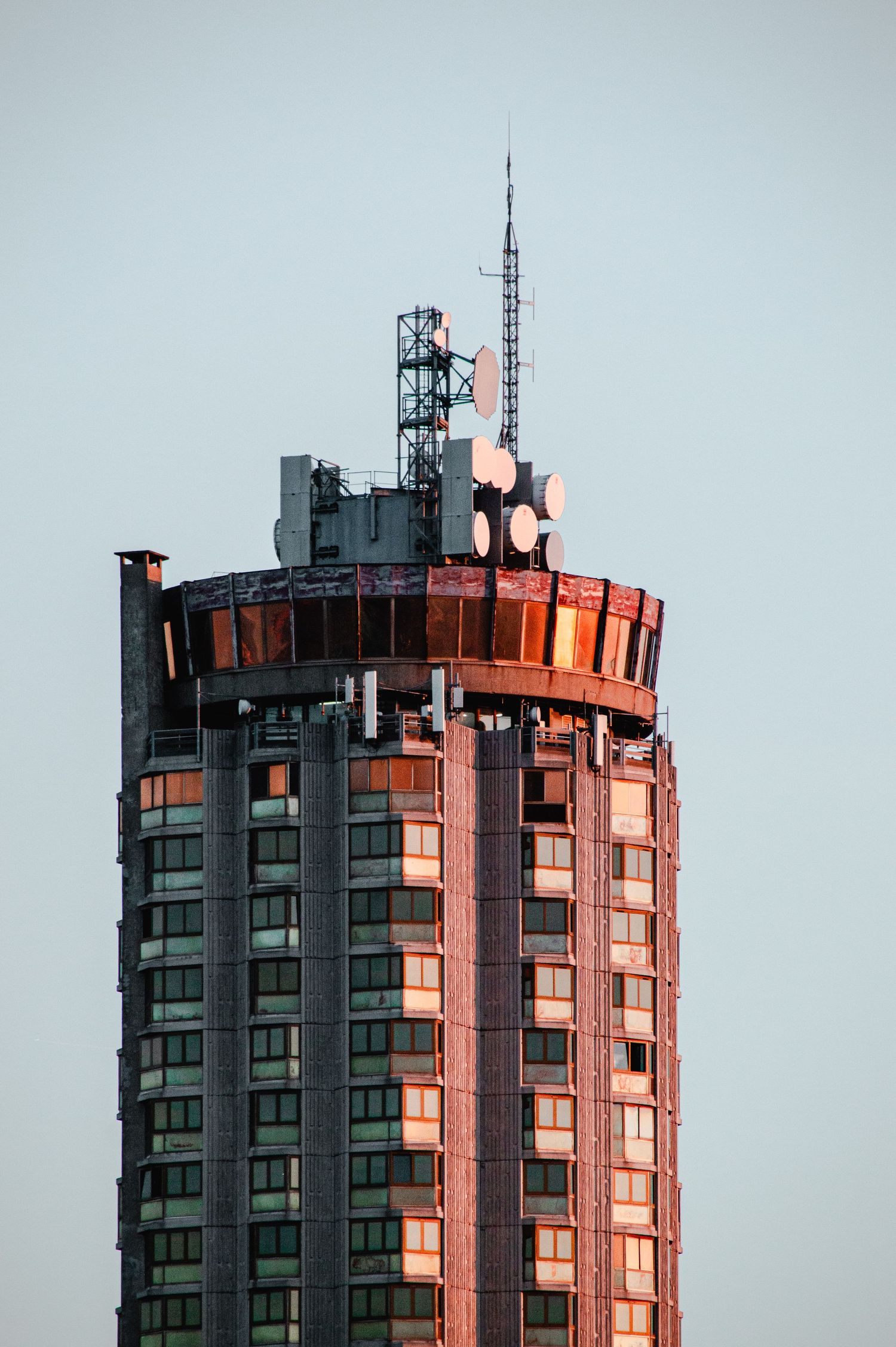 Tall cylindrical building with red-tinted windows reflecting sunset light and communication towers on its roof.