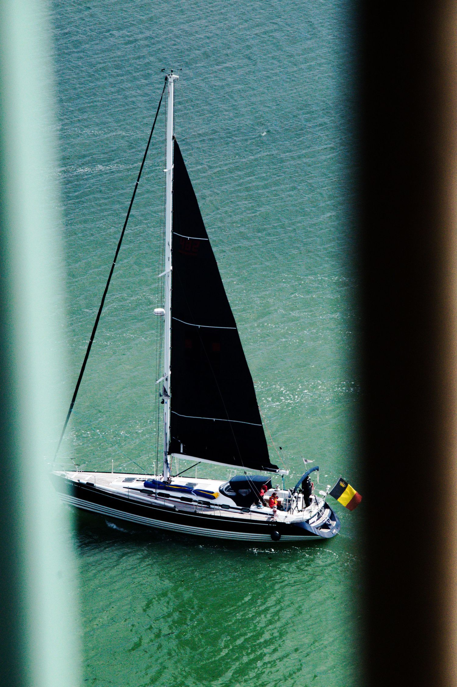 Aerial view of a sailboat with black sails flying a Belgian flag on turquoise water, framed by a bridge structure.