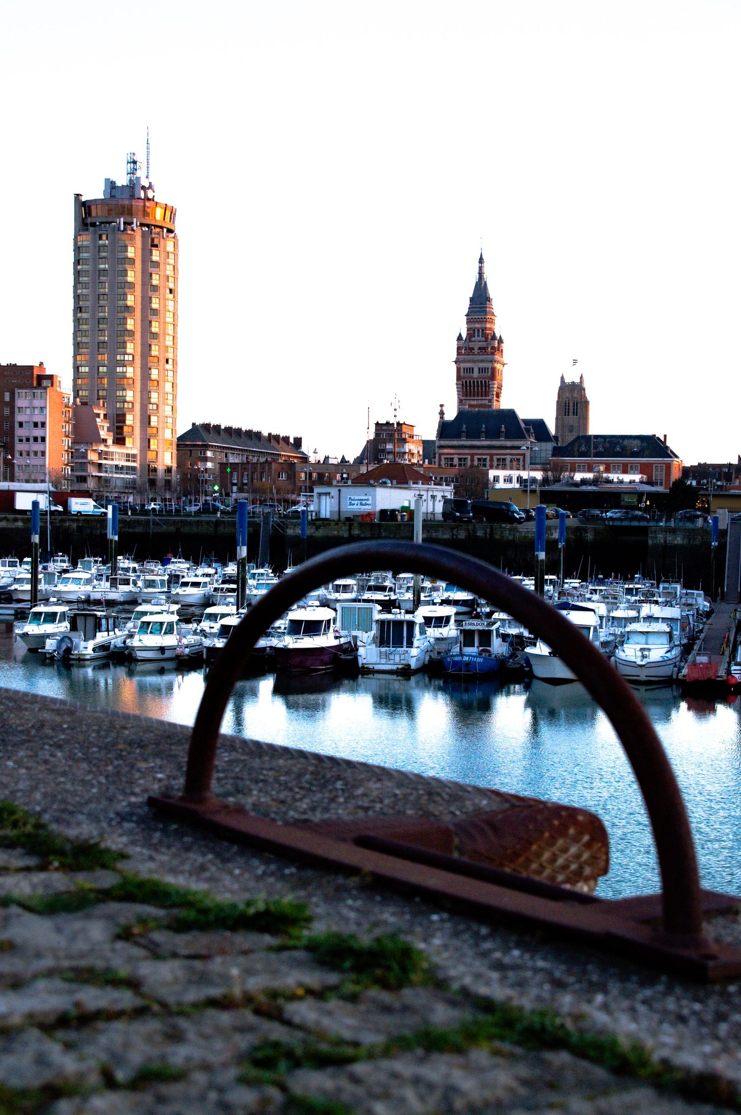 Marina at dusk with boats and city buildings silhouetted against a darkening sky in Dunkerque, France.