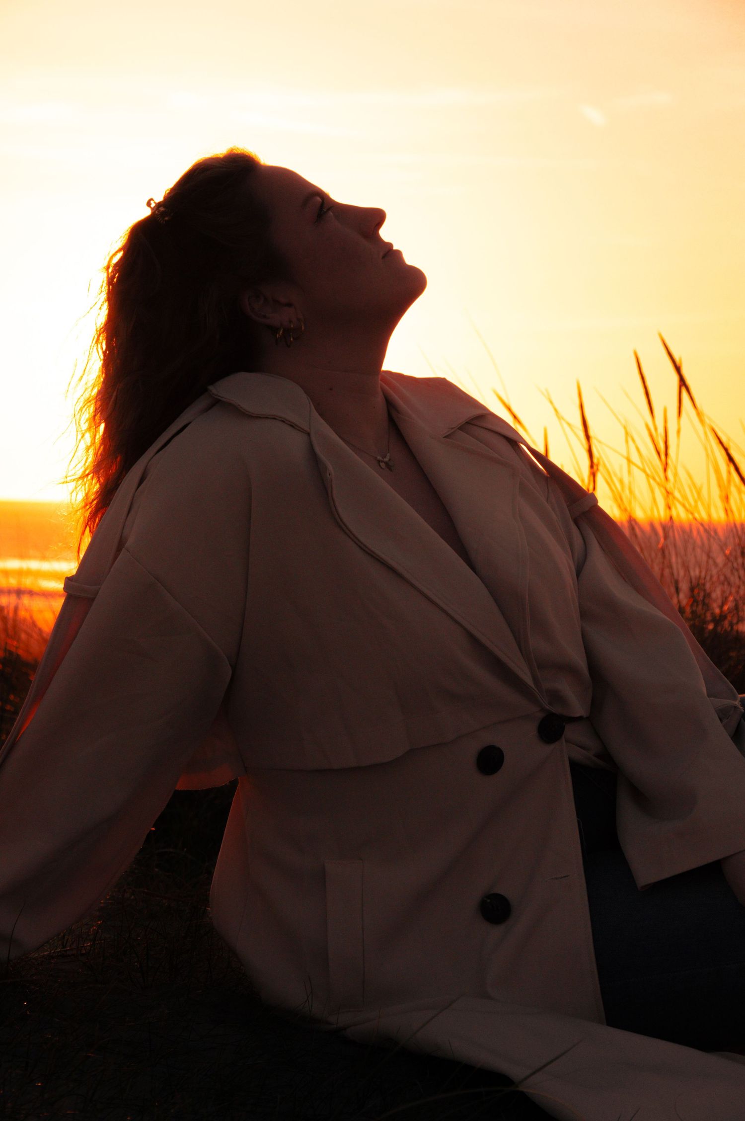 Woman in beige trench coat sits outdoors, head tilted back against golden sunset sky.