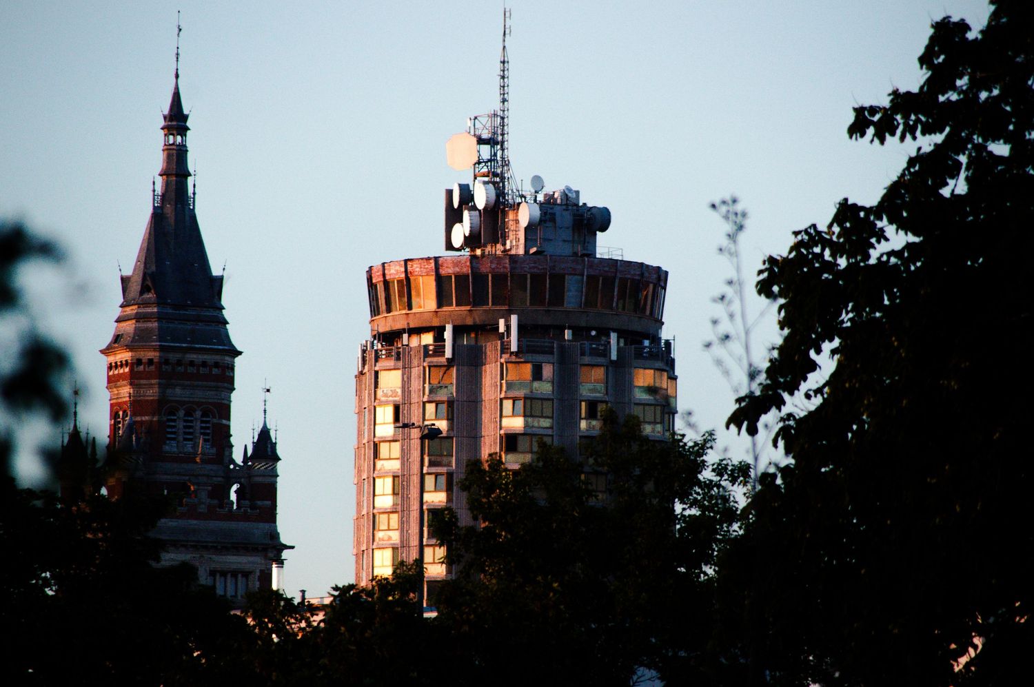 Cylindrical tower and Gothic church spire rise above silhouetted trees at dusk in an urban skyline.