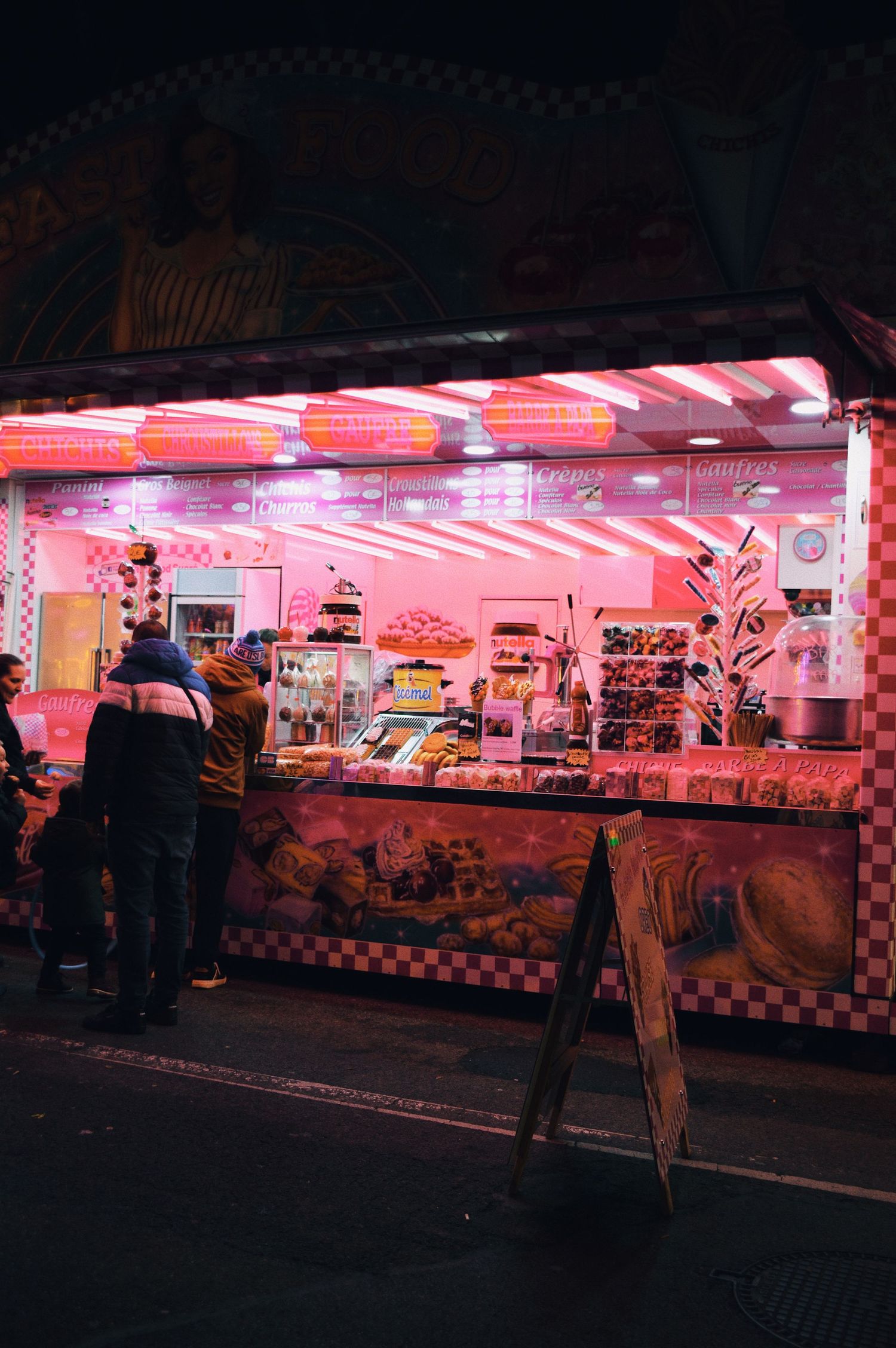Pink-lit carnival food stand with retro diner styling and customers waiting in line.