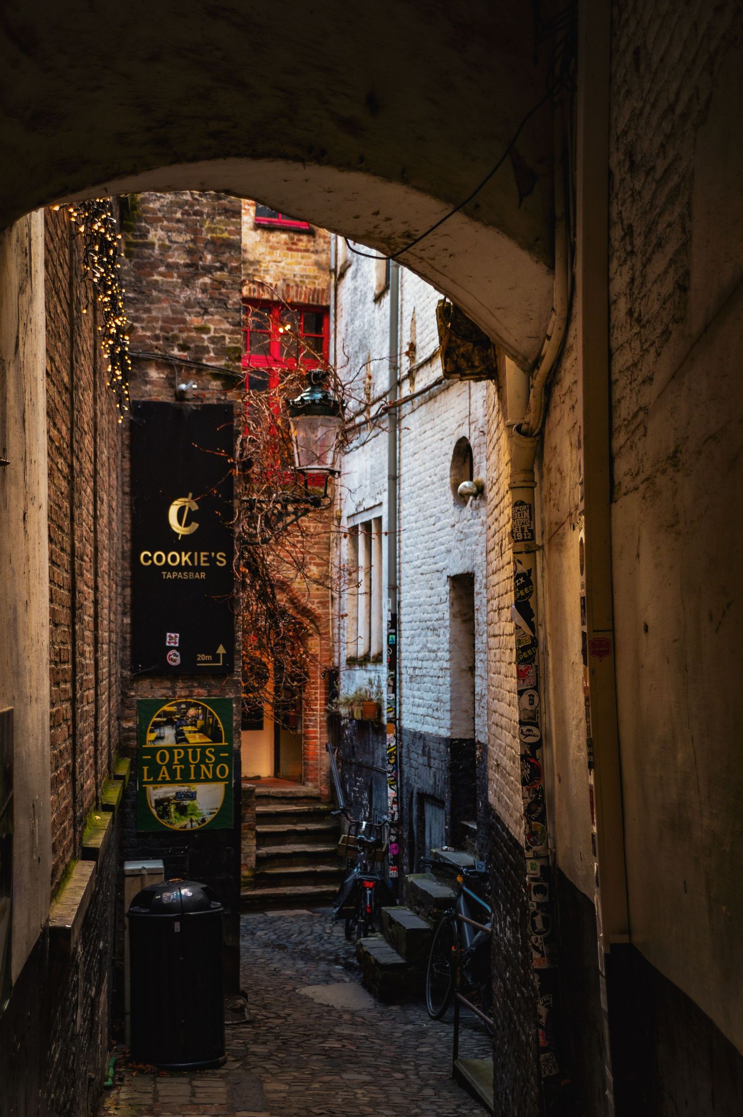 Narrow medieval alley with arched passageways and dimly lit shop signs.