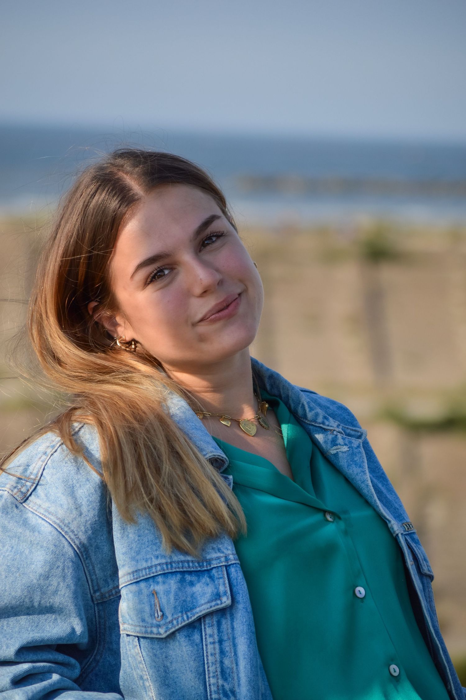 Woman in denim jacket smiling against coastal background on sunny day.