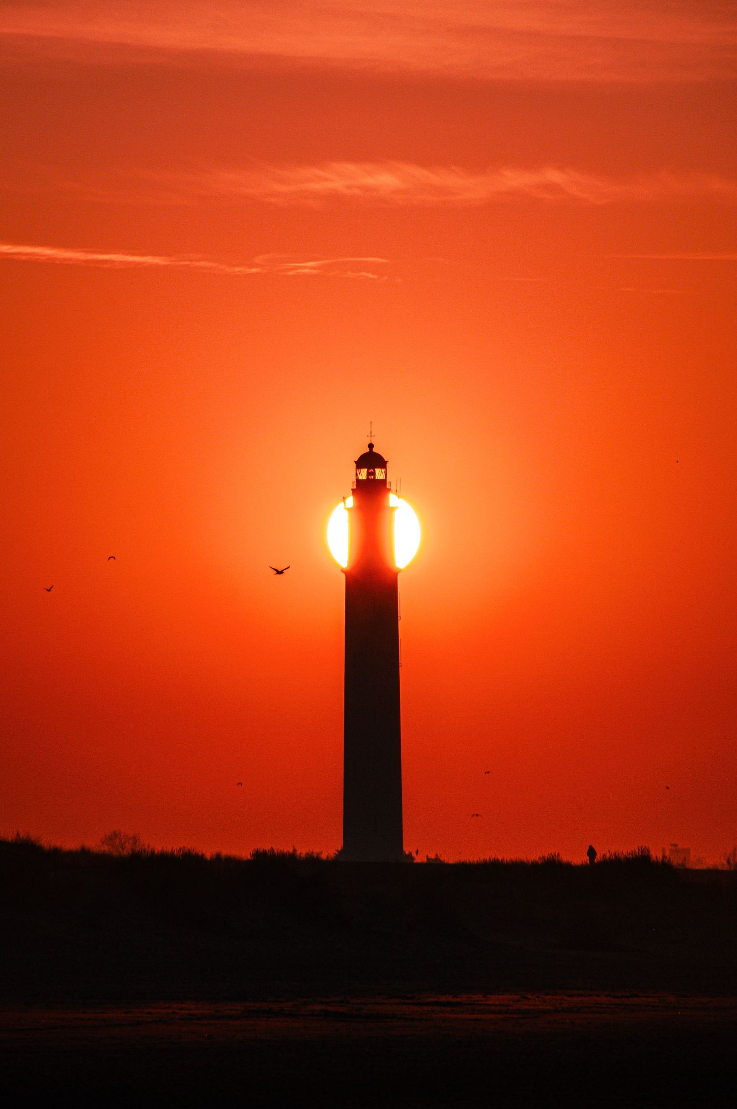 Silhouette d’un phare sur fond de ciel spectaculaire de coucher de soleil orange, avec le soleil parfaitement aligné derrière la tour.
