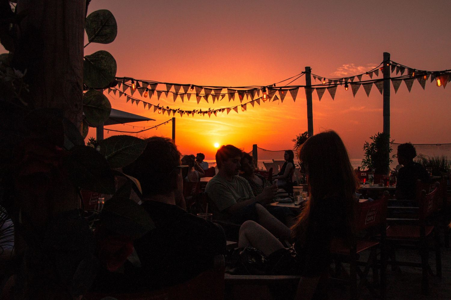 Silhouetted people dining at an outdoor rooftop bar with string pennant lights against a vibrant orange sunset sky.