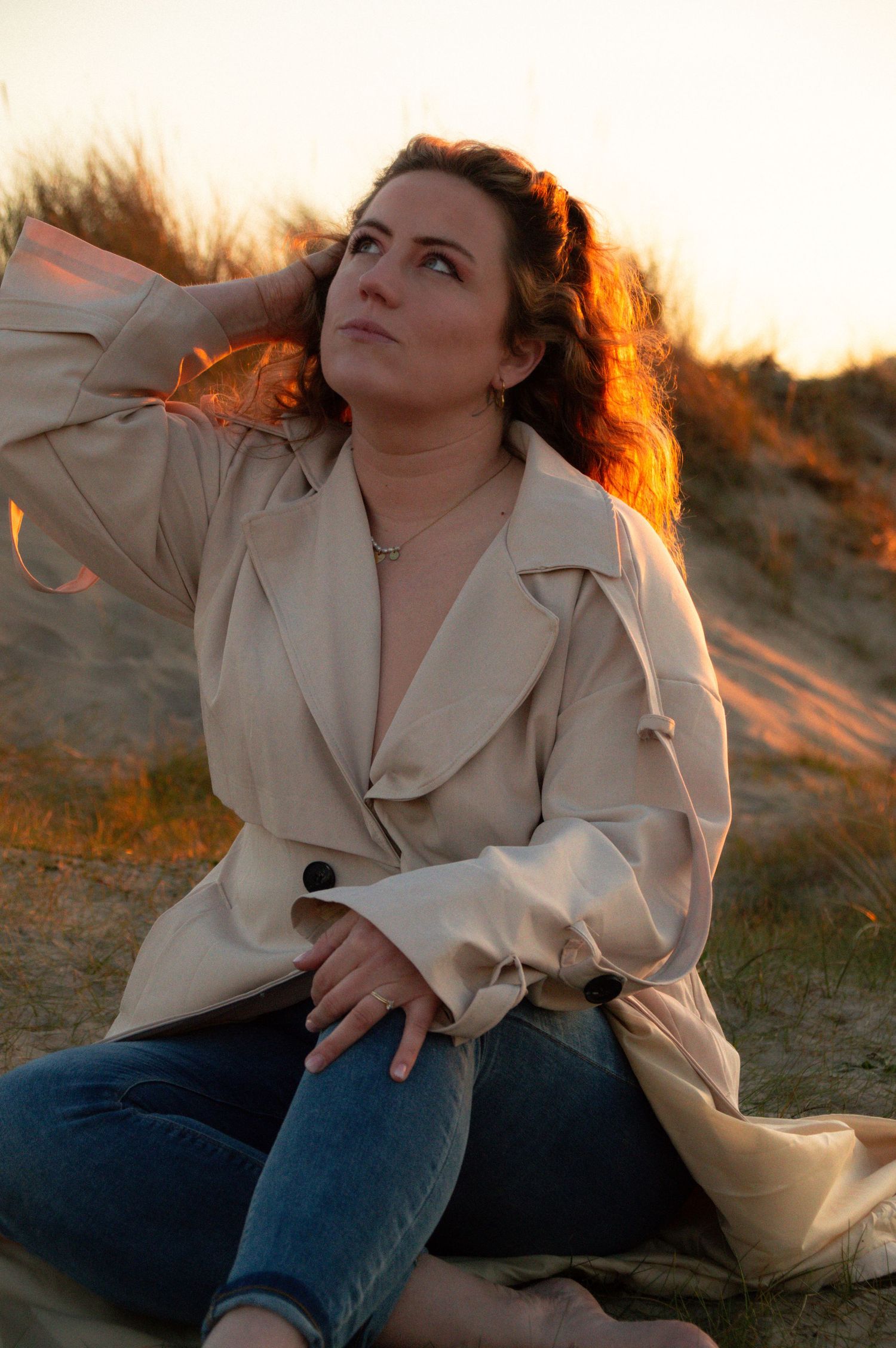 Woman in beige trench coat and jeans sitting on sandy dunes at golden sunset, gazing upward thoughtfully.