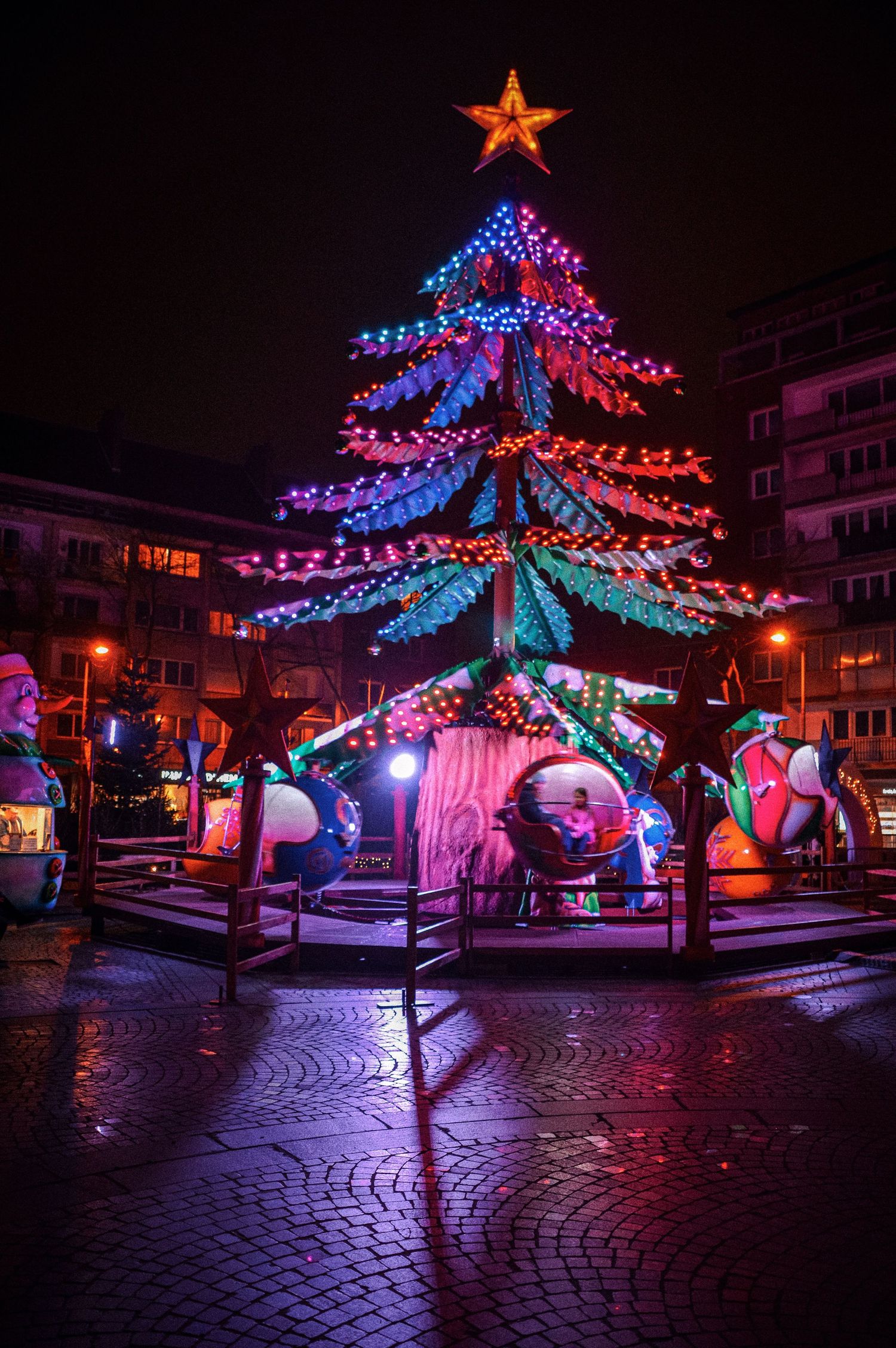 Large illuminated Christmas tree display with red and blue lights in a town square at night.