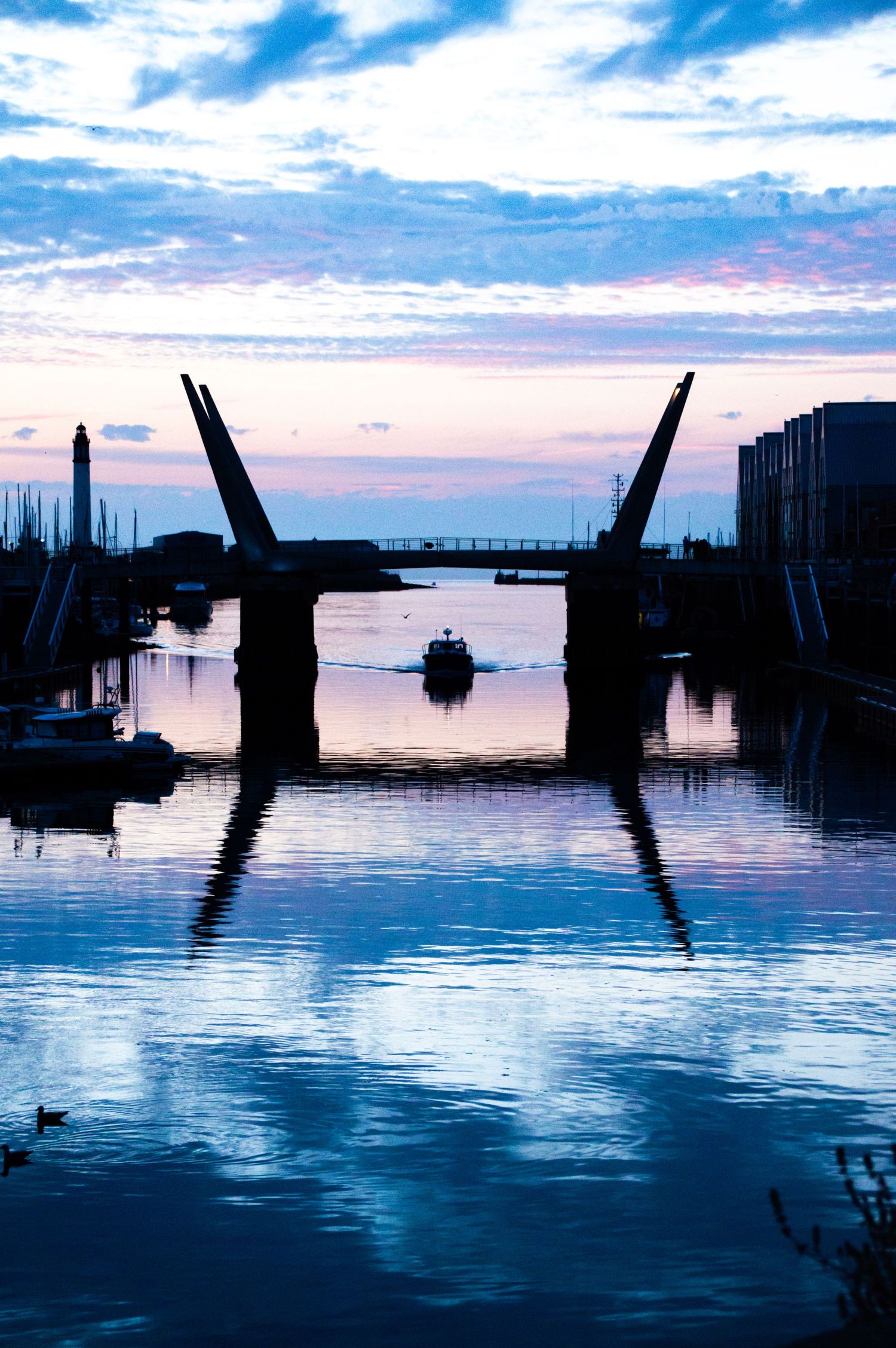 Silhouette of a drawbridge at dusk with a boat passing through calm reflective waters under a blue and pink sunset sky.