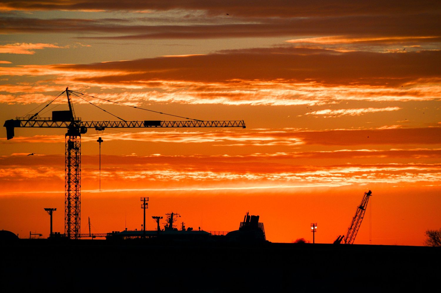 Silhouette of construction cranes against a dramatic orange sunset sky.