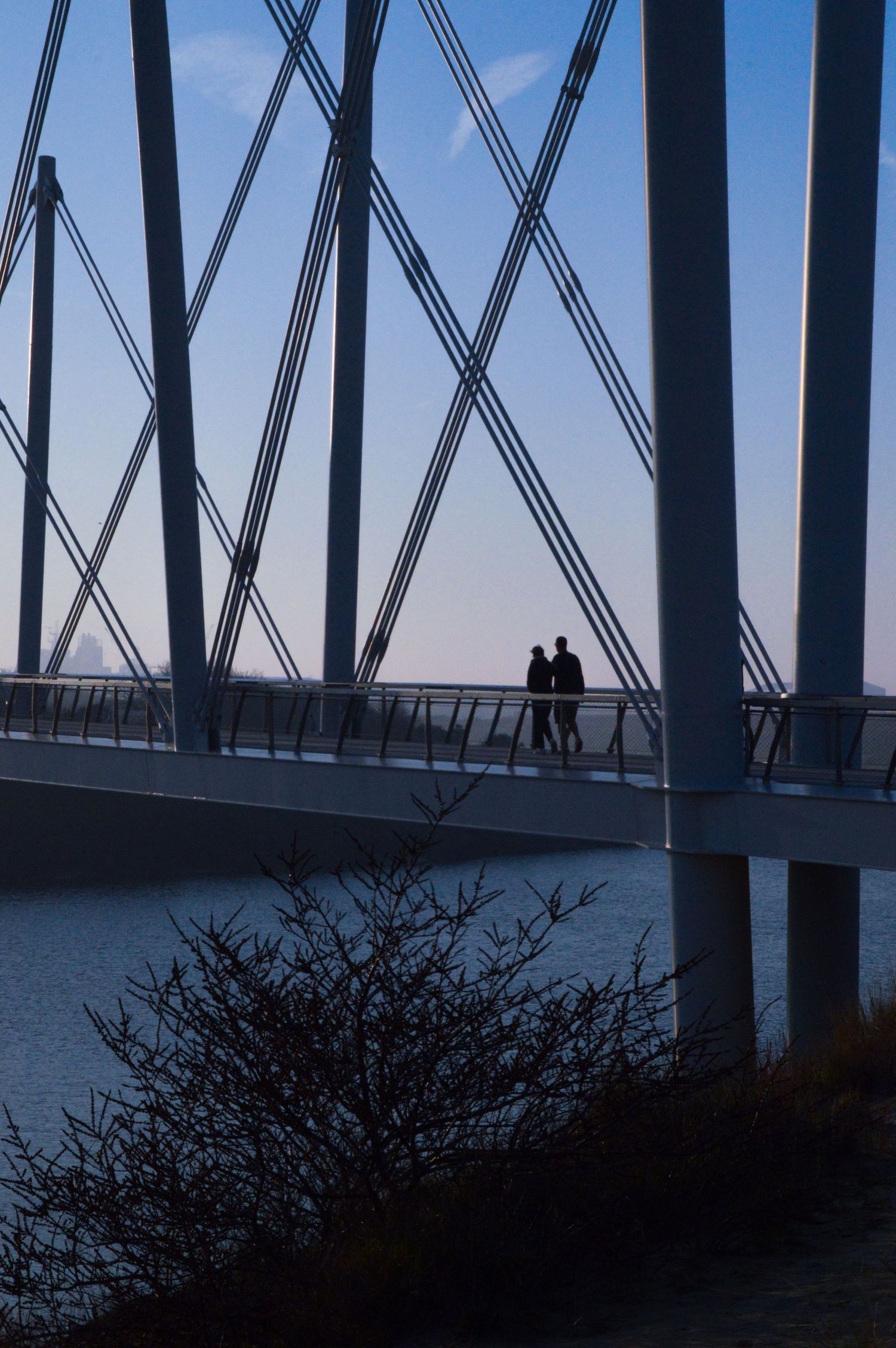Deux silhouettes sur une passerelle de pont encadrée par des poutres métalliques industrielles en croix, sur fond de ciel bleu.
