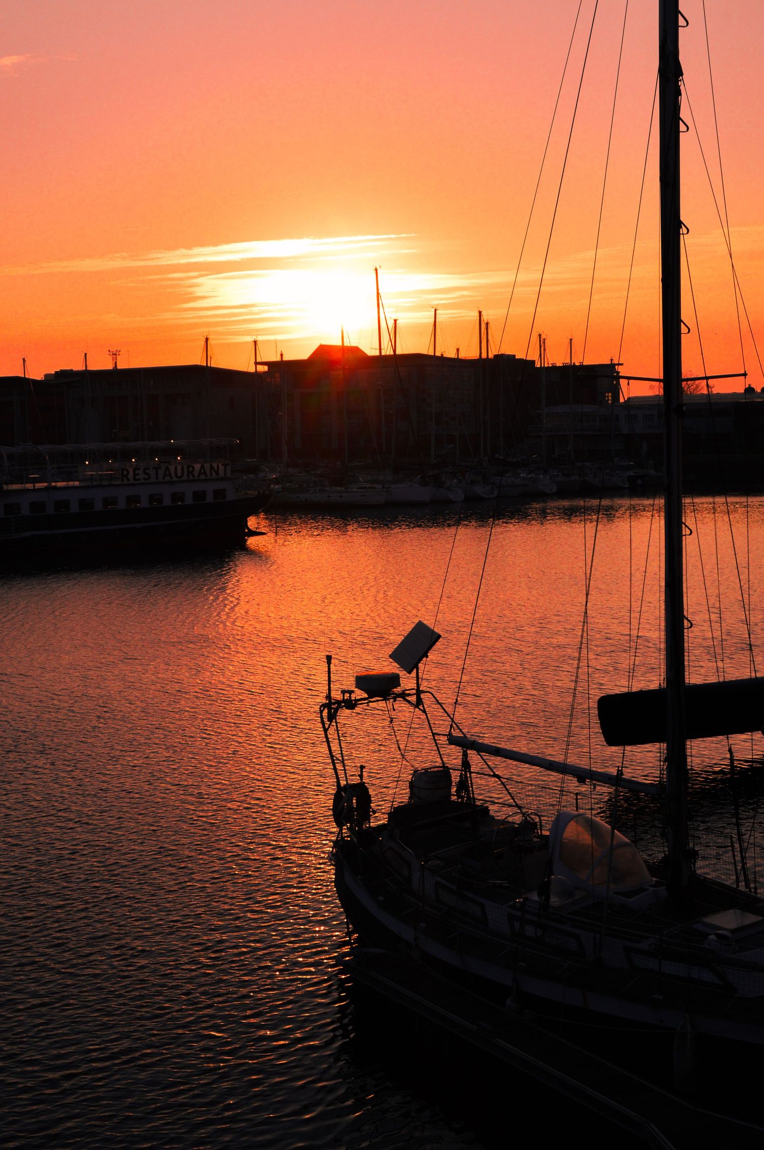 Sailboat silhouetted against vibrant orange sunset at marina harbor with masts and rigging visible.