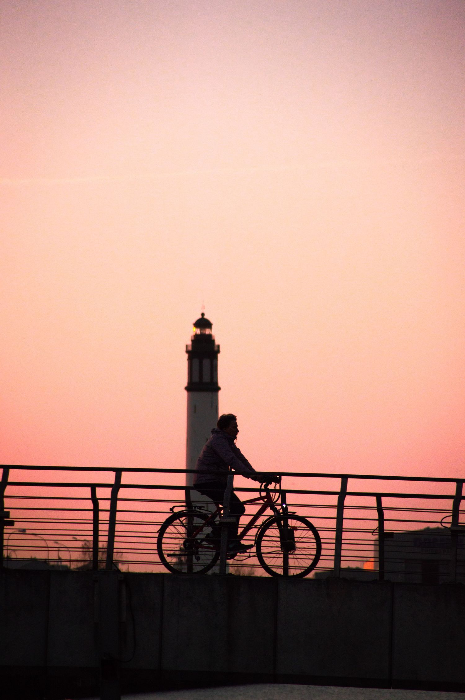 Silhouette of a lighthouse and bicycles on a railing against a pink sunset sky.