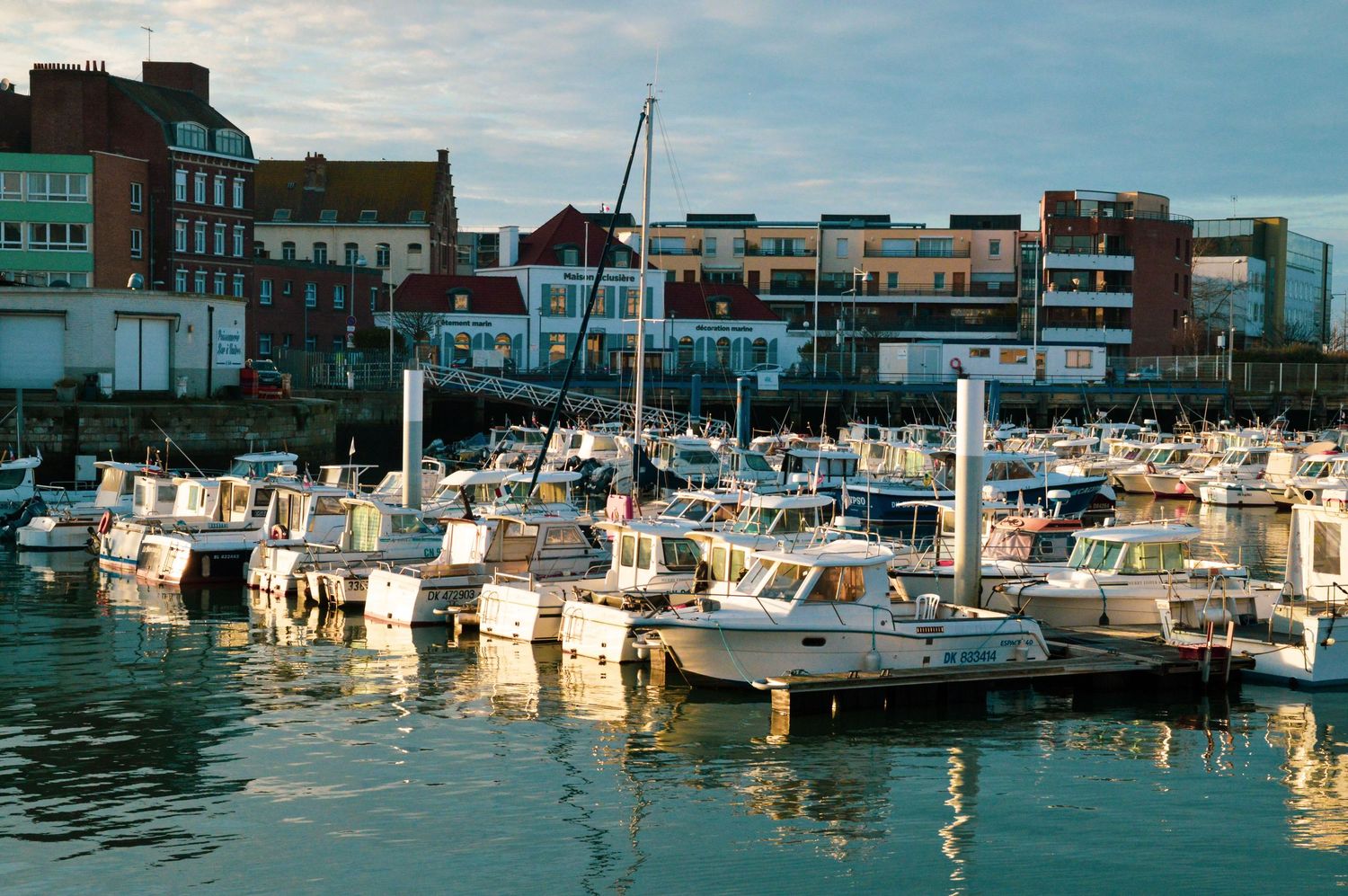 Marina with boats docked at a waterfront harbor on a sunny day with buildings in the background.