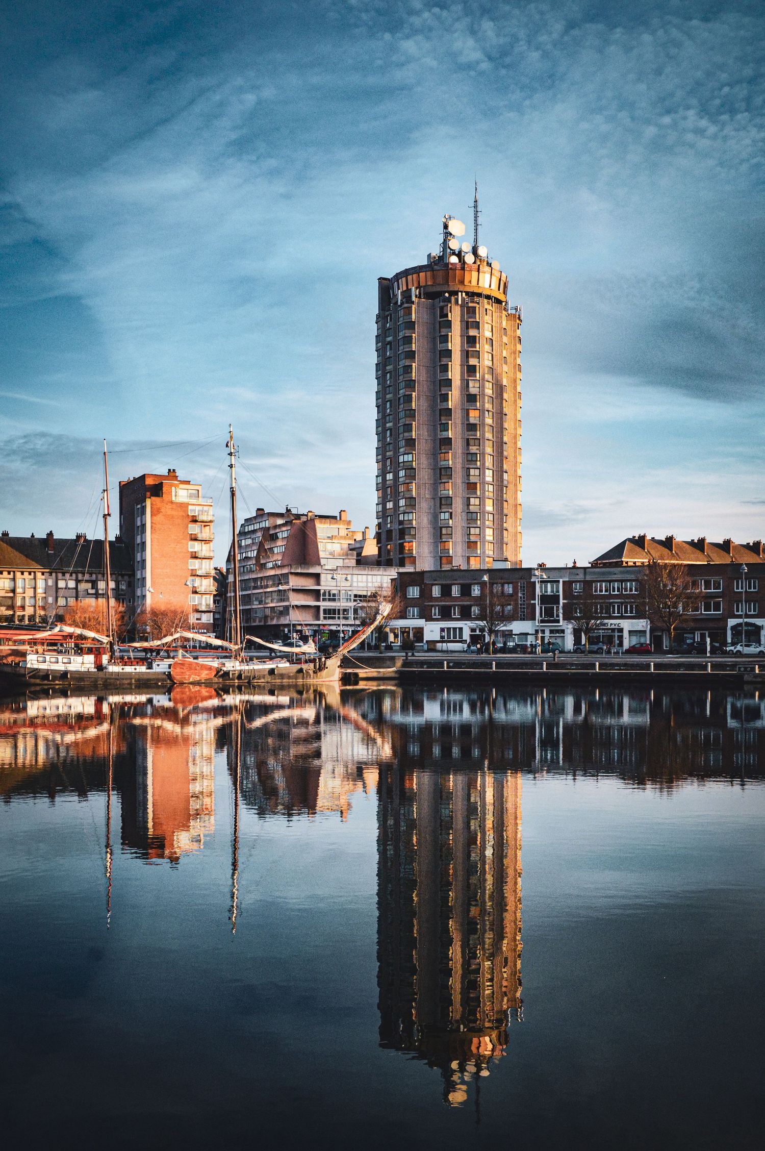 Modern apartment buildings reflecting in calm harbor waters during sunset.