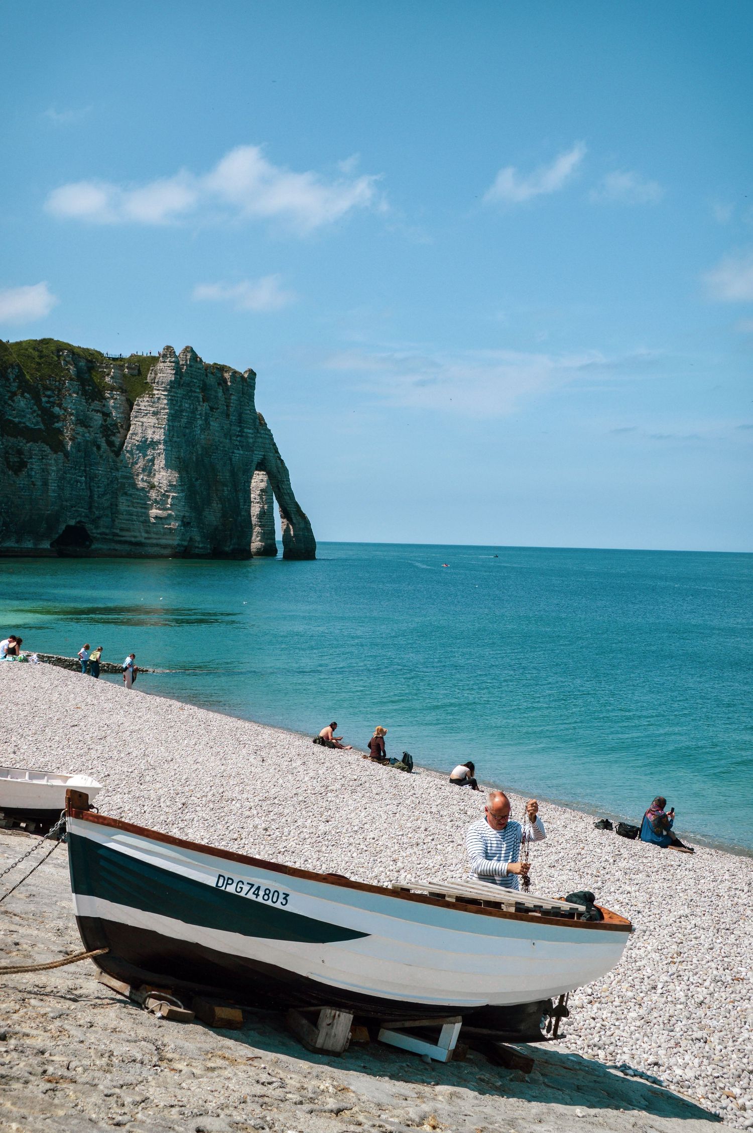 Beachgoers relax near a boat on a pebble beach beneath towering limestone cliffs in Etretat.