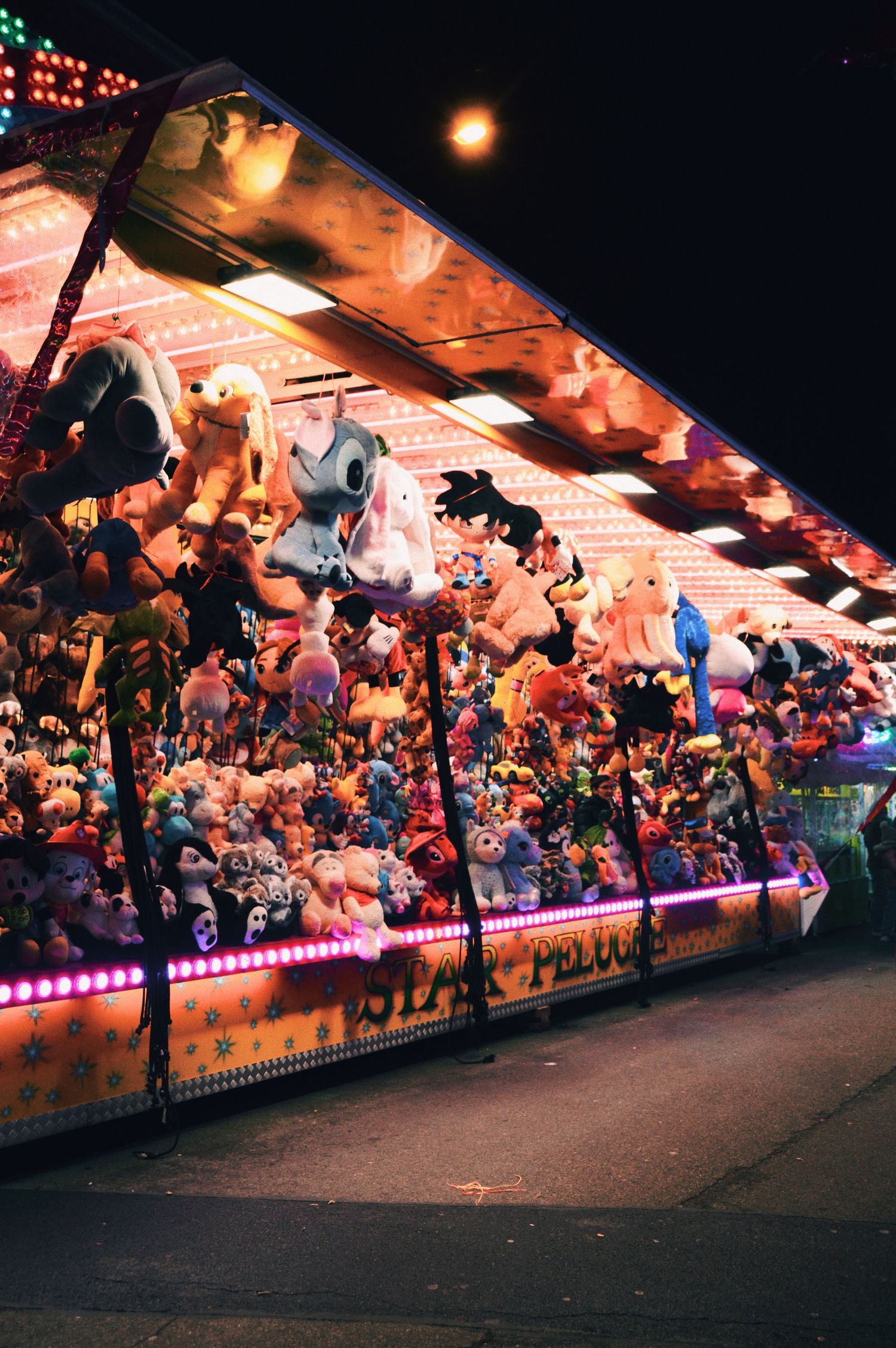 Long row of carnival game stalls displaying colorful stuffed animal prizes under bright lights.