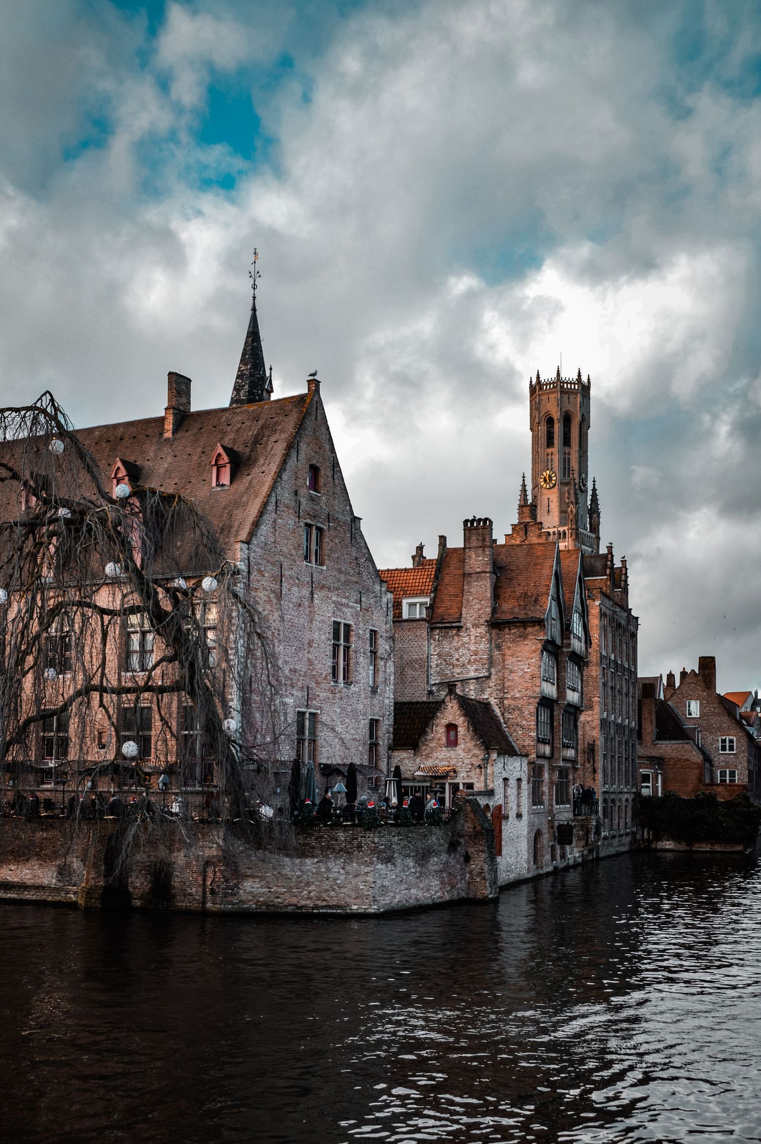 Medieval buildings and church tower along a canal in Bruges with dramatic cloudy sky.