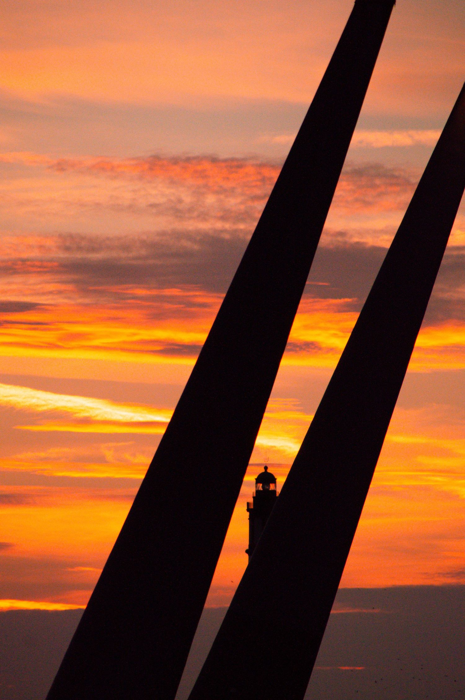 Silhouette of a lighthouse framed between two diagonal structures against a vibrant orange and red sunset sky.