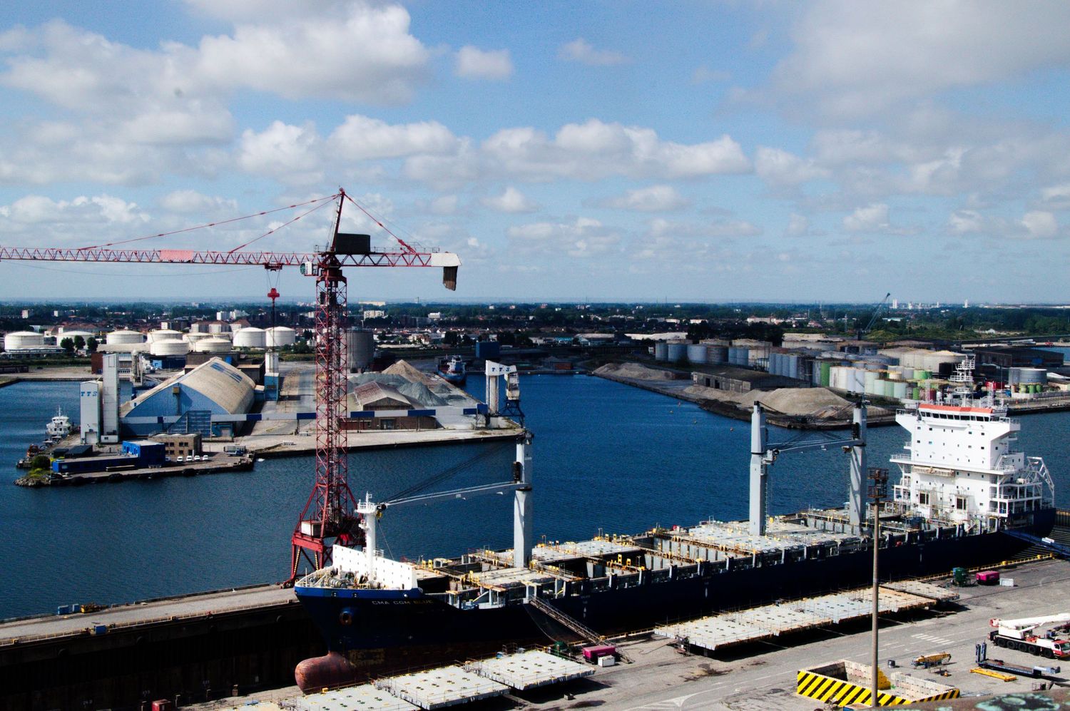 Aerial view of a busy industrial harbor with a red crane, docked cargo ships, and storage facilities under a cloudy sky.