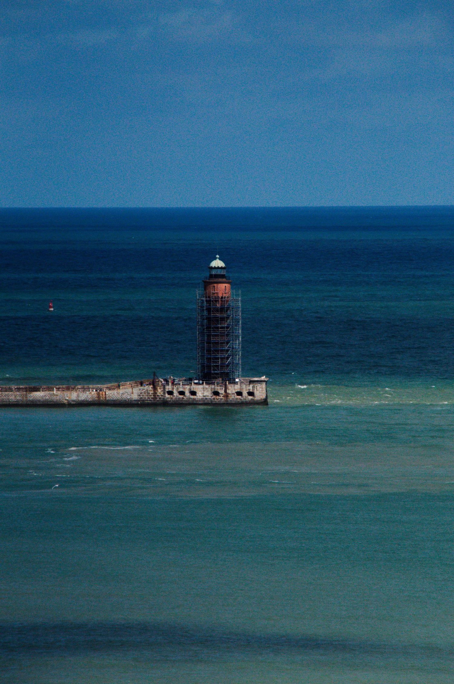 A lighthouse under scaffolding renovation stands on a concrete pier surrounded by turquoise and deep blue ocean waters.