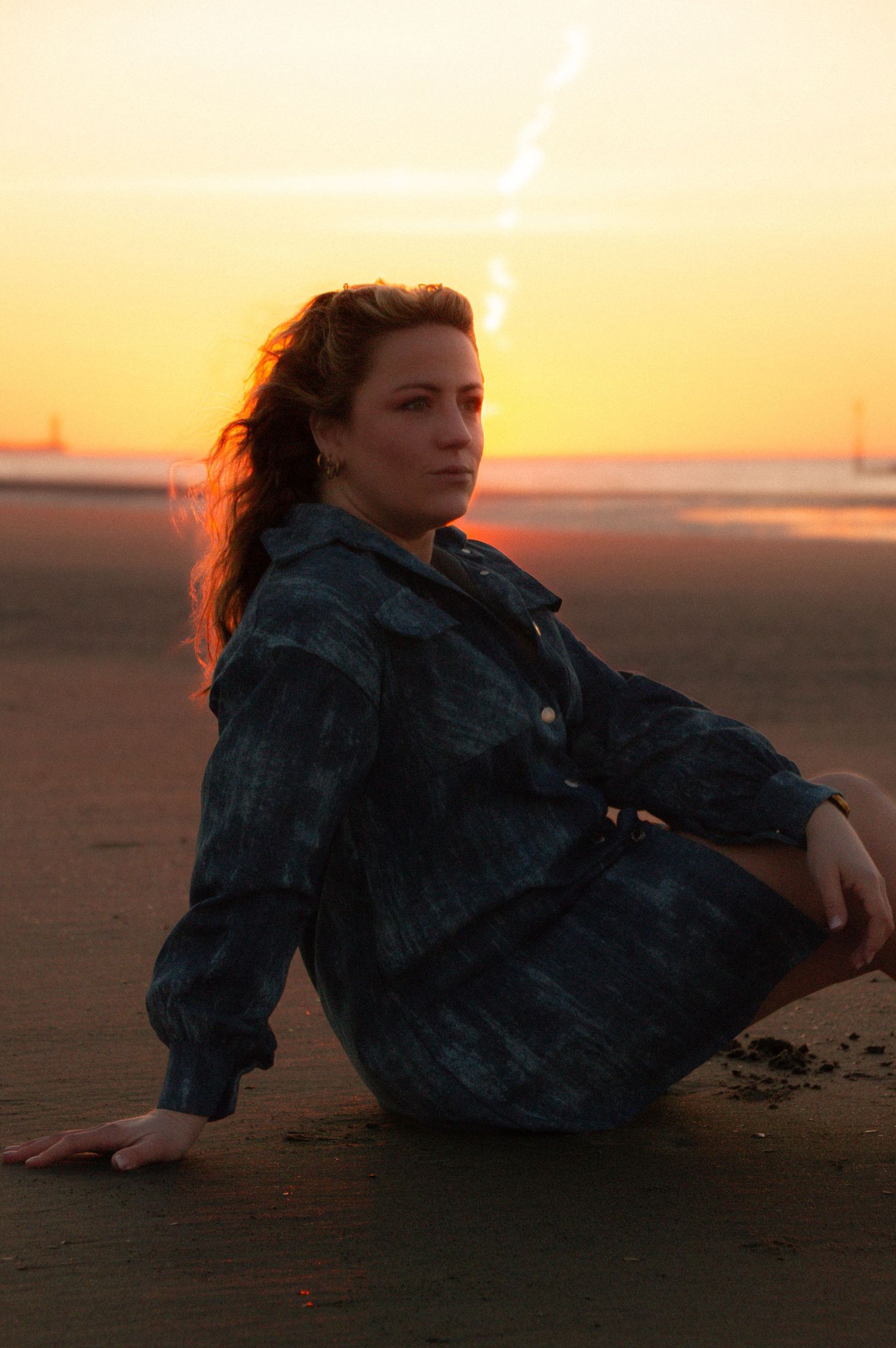 Woman in dark outfit sitting on beach sand during golden sunset with warm orange sky backdrop.