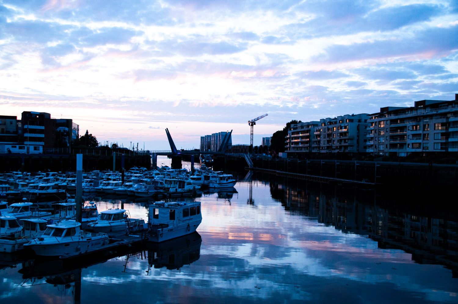 Tranquil marina at dusk with boats reflecting on calm water, city buildings and crane silhouetted against a blue cloudy sky.