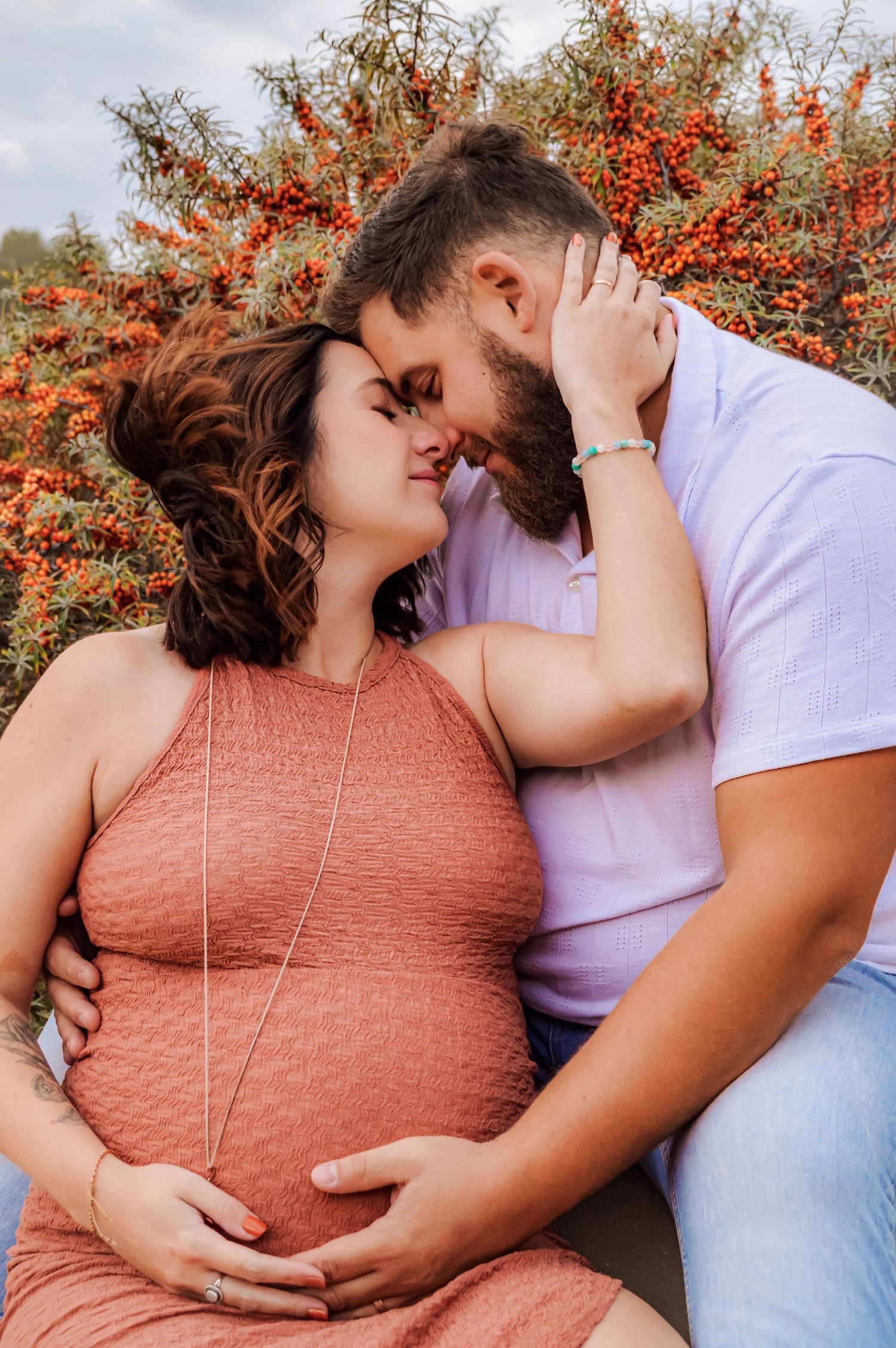 Couple embracing during outdoor maternity session with red berries in background.