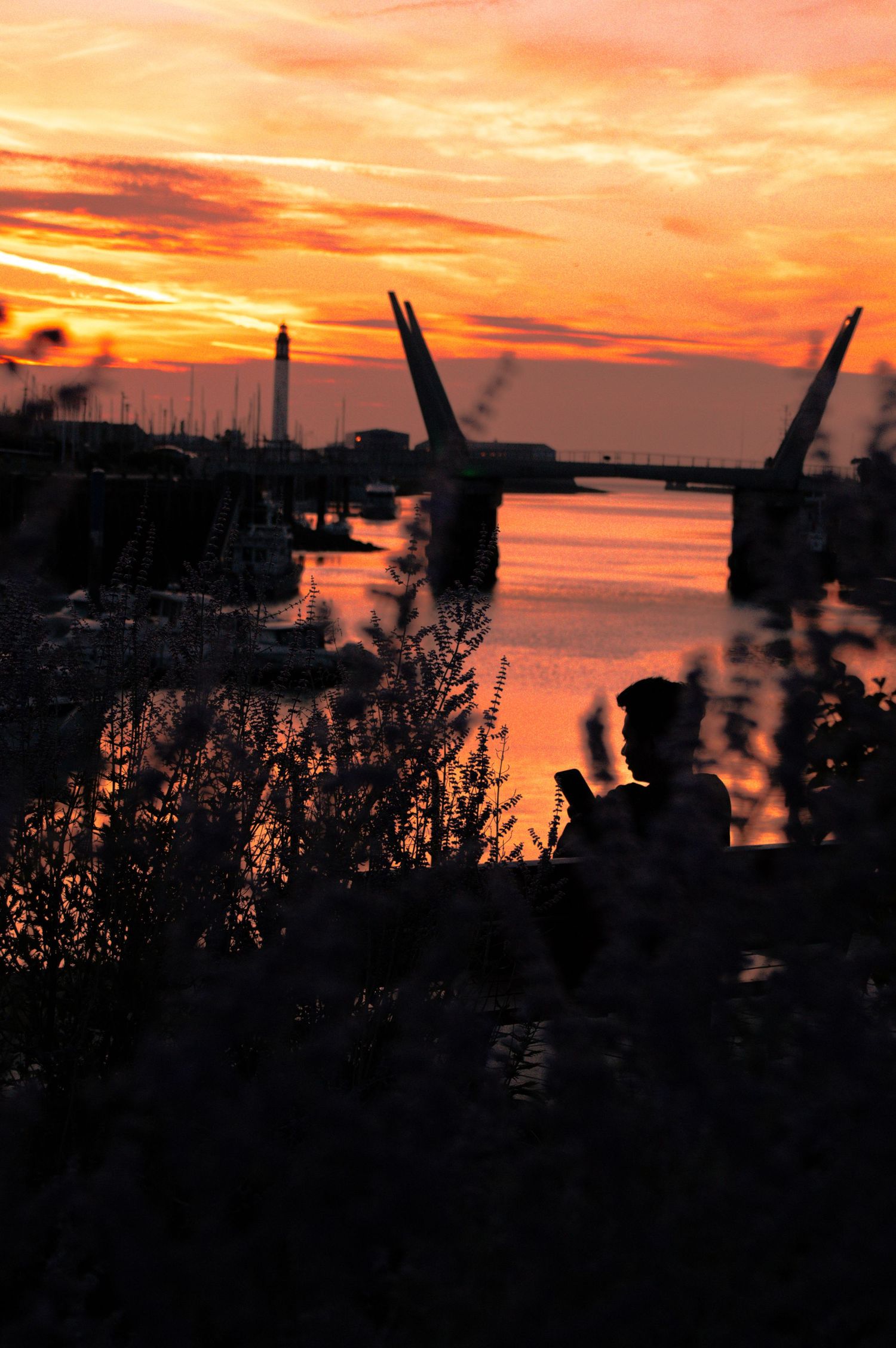 Silhouette of person on phone at sunset by a harbor with a drawbridge and vivid orange sky reflecting on water.