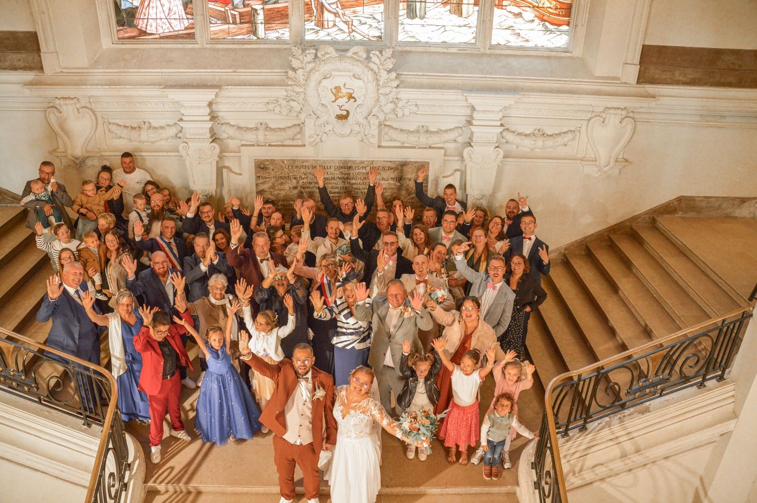 Large group wedding photo on grand staircase in ornate interior.