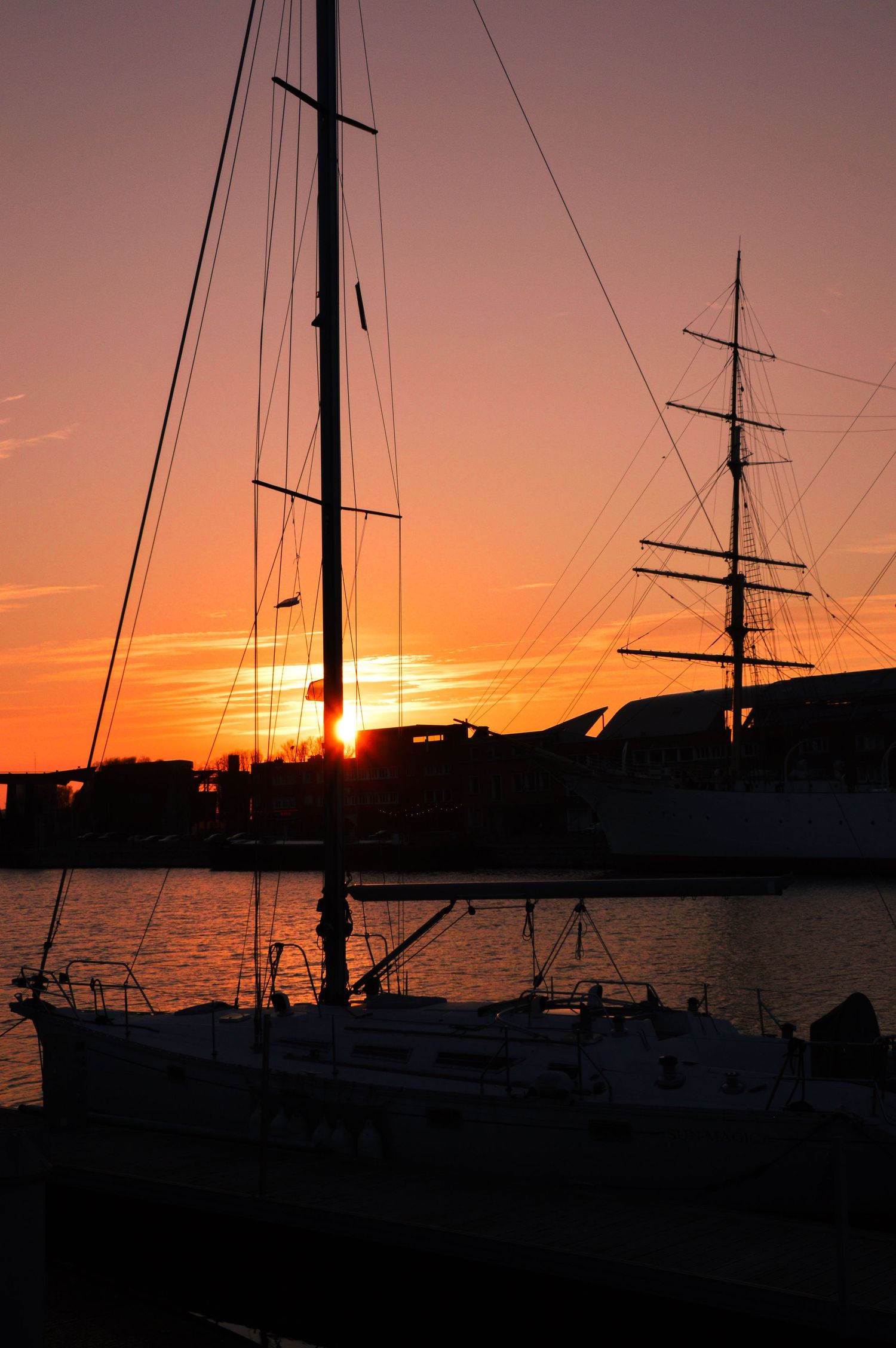 Two sailing vessels in harbor silhouetted against dramatic orange sunset sky.