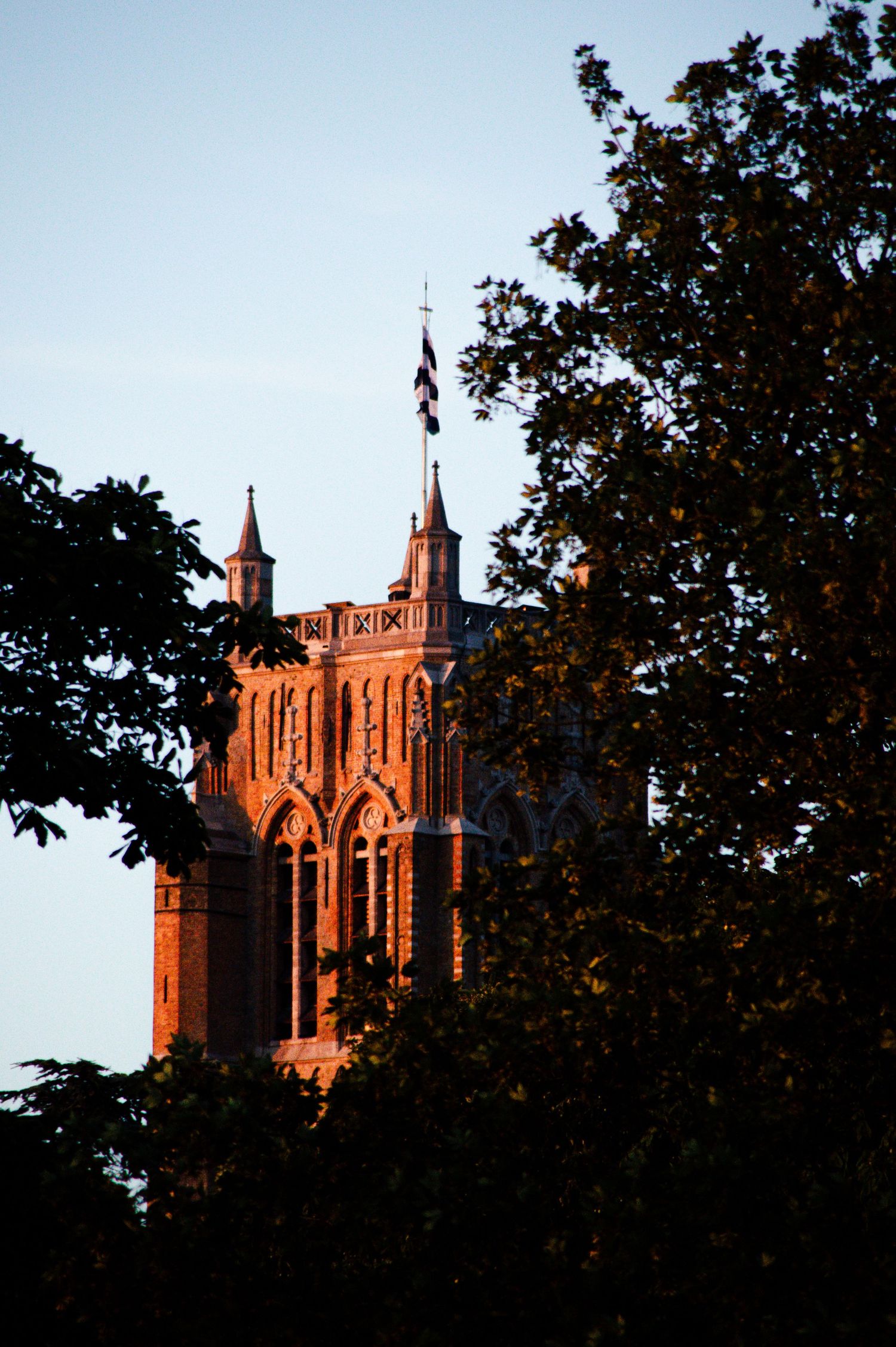 Gothic red brick church tower with a flag at sunset, framed by dark silhouetted trees against a blue sky.
