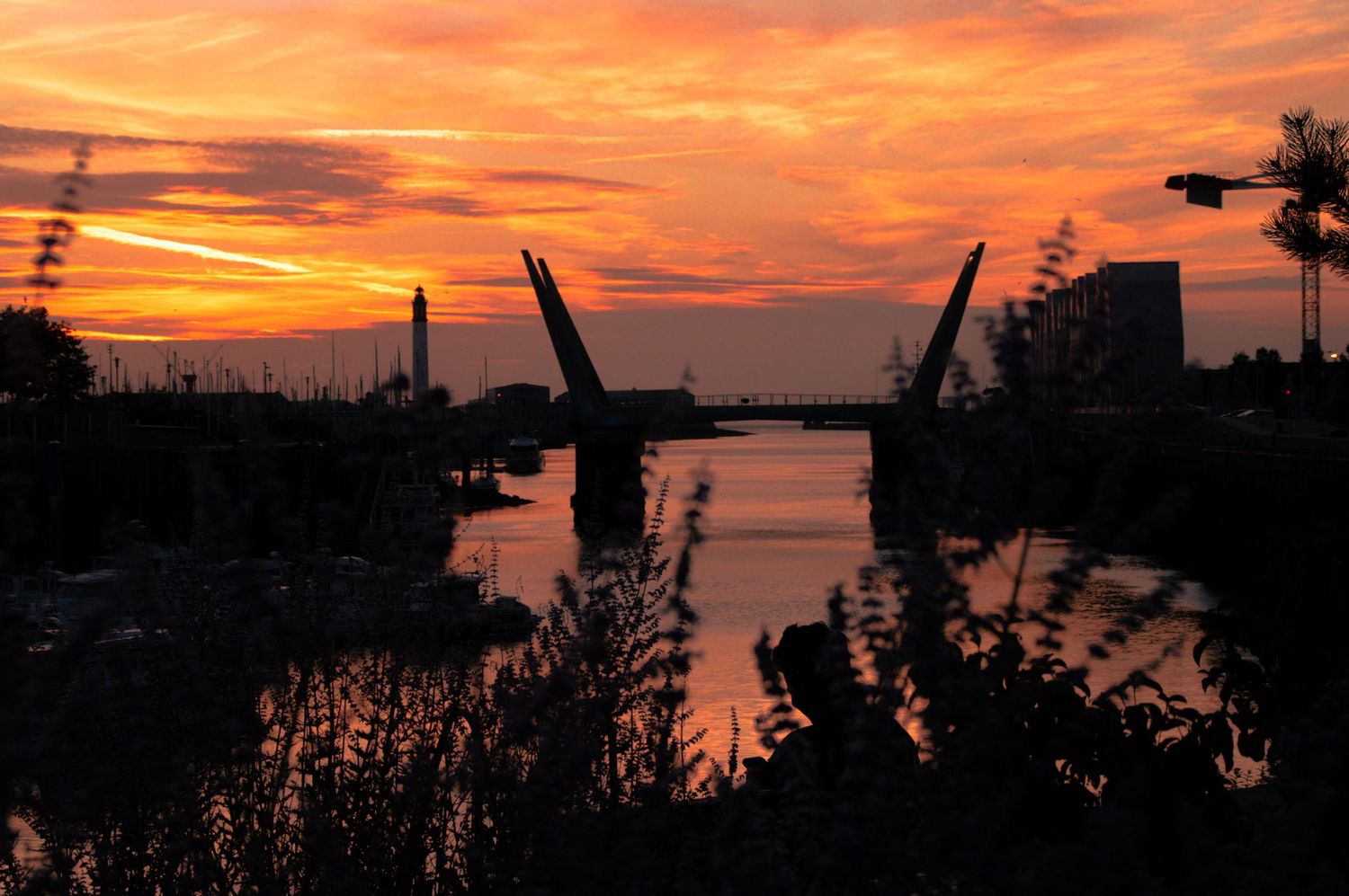 Stunning orange and red sunset silhouettes a drawbridge and lighthouse over a calm river channel.