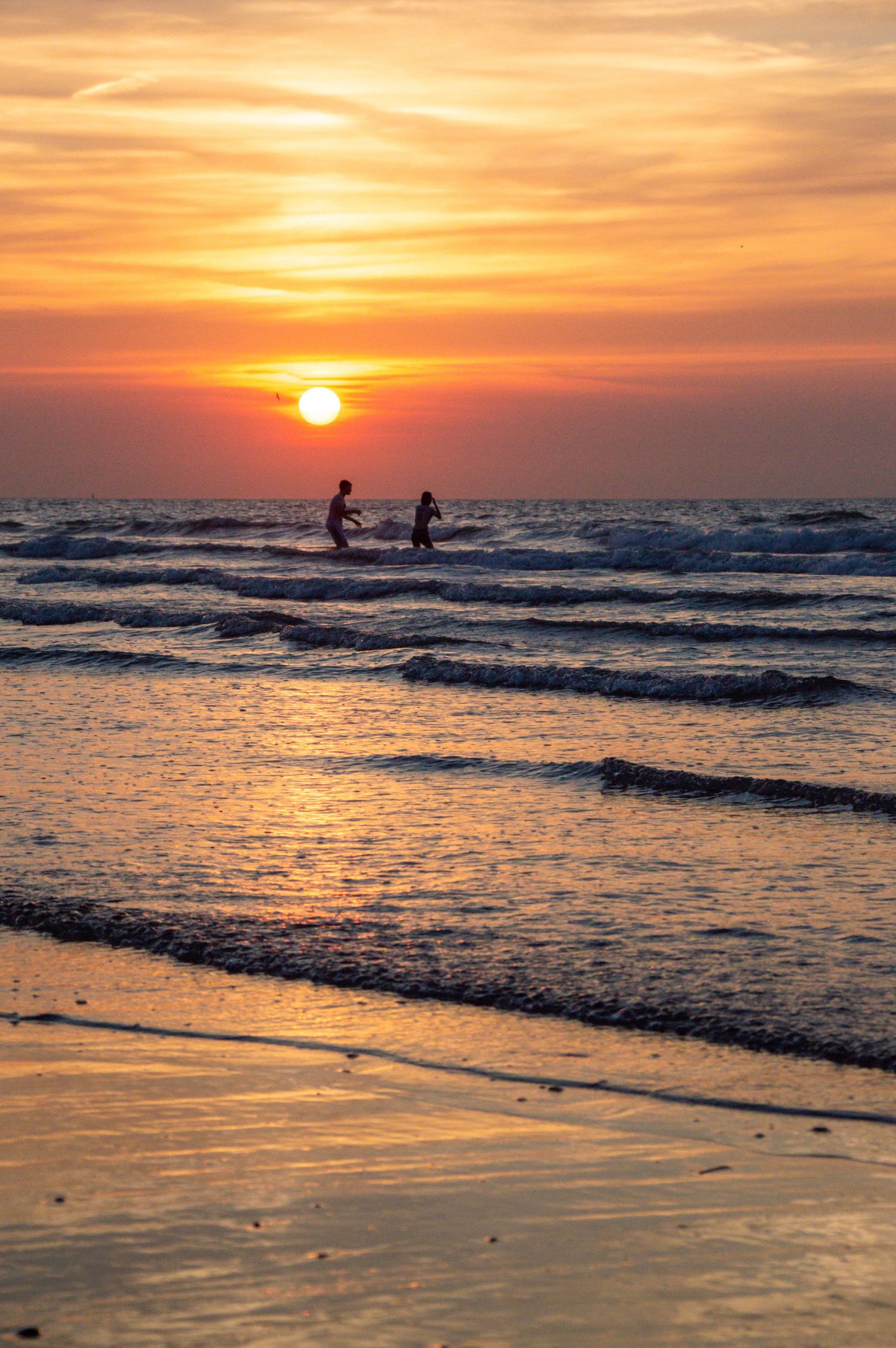 Two silhouetted figures wade in ocean waves during a stunning golden sunset at the beach.