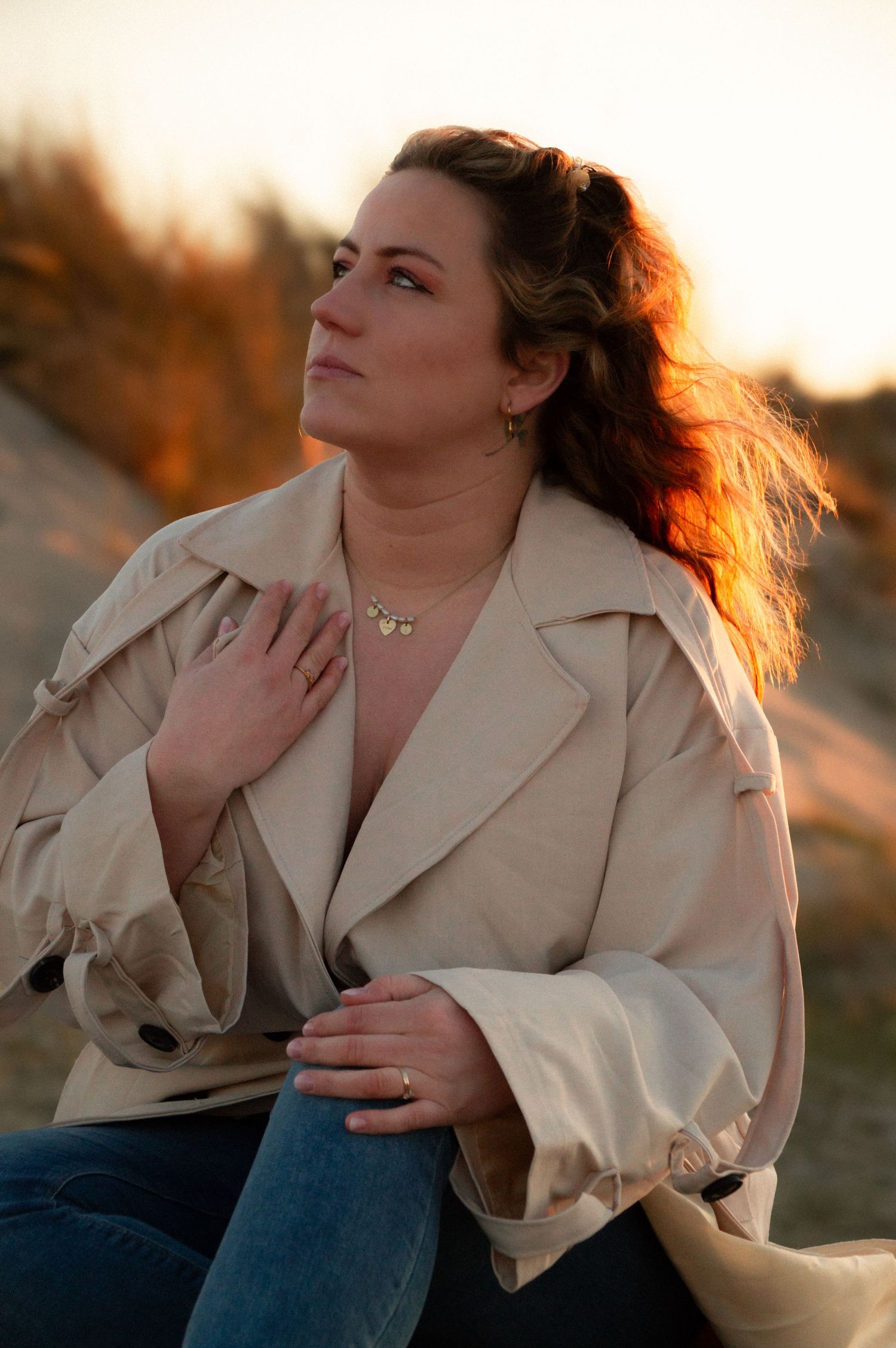 Woman in beige trench coat sits outdoors at sunset, hand on chest, looking thoughtfully away.
