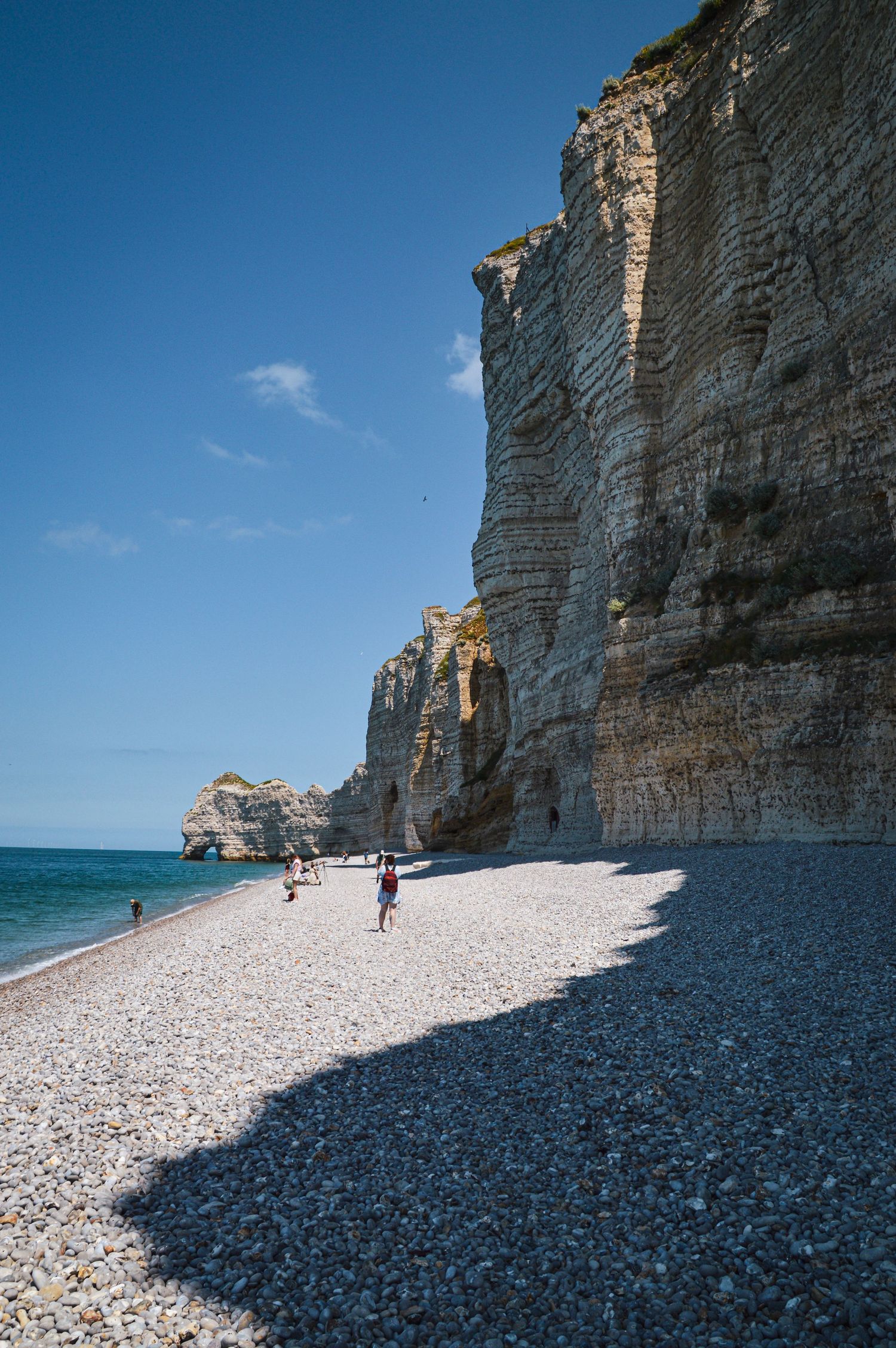 A pebble beach stretches along massive limestone cliffs under bright blue skies in Normandy.