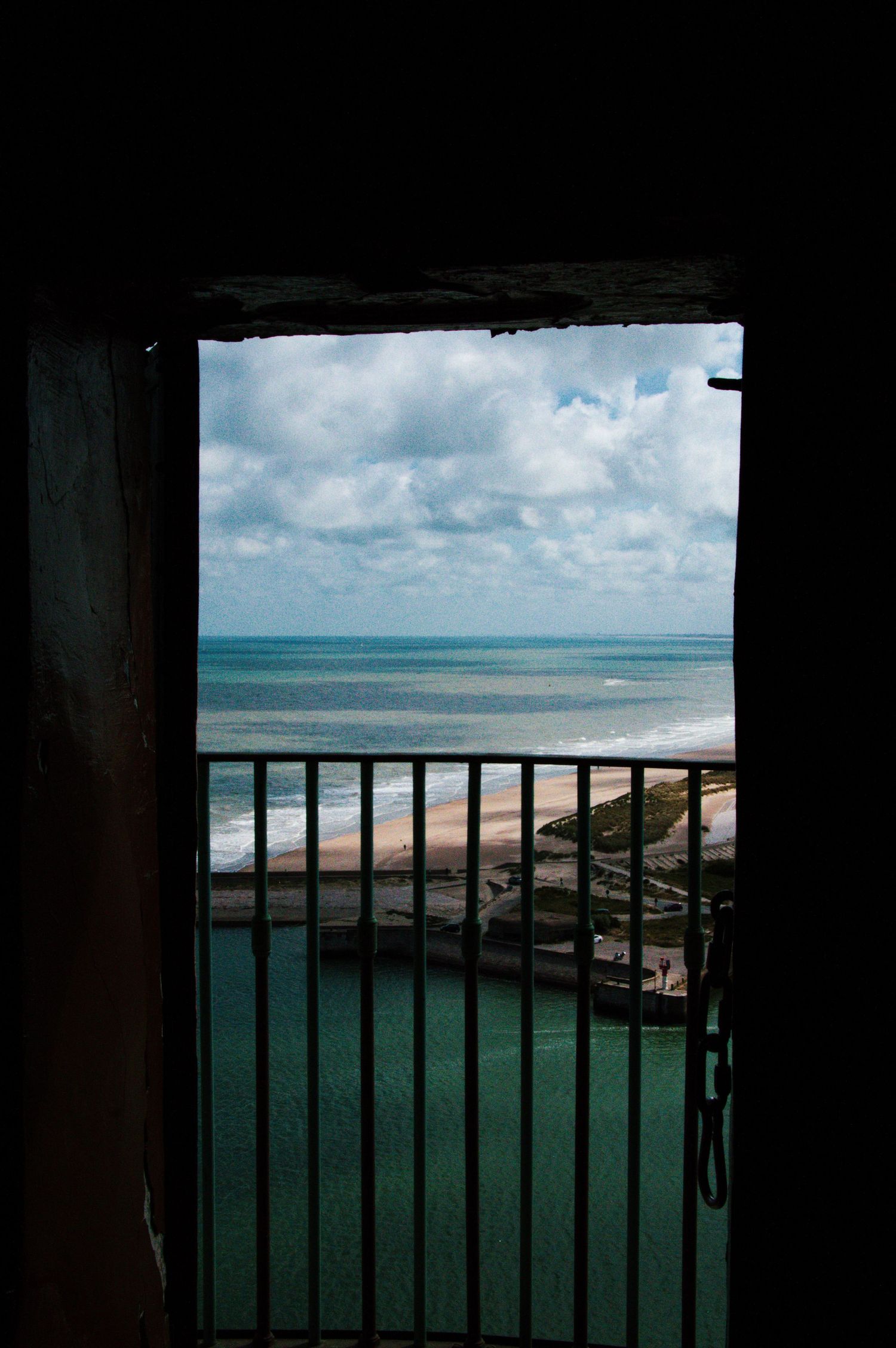 View through a dark doorway onto a balcony with iron railings overlooking a sandy beach and turquoise ocean.