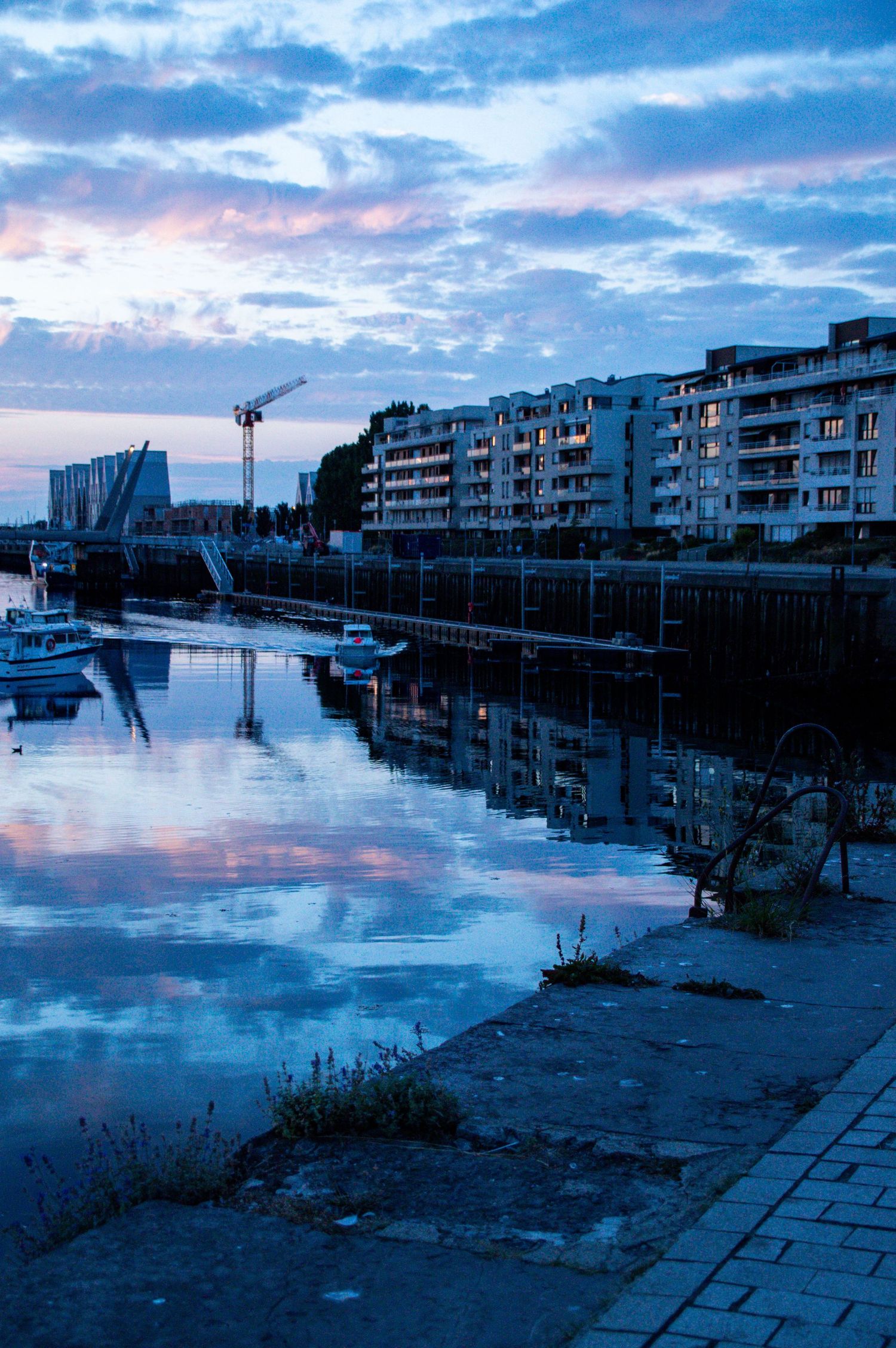 Blue hour cityscape reflecting on calm harbor waters with apartment buildings, boats, and a construction crane at dusk.