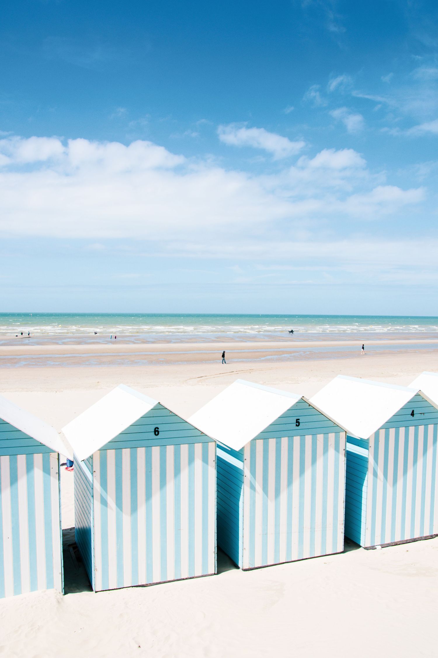 Row of blue and white striped beach huts on a sandy beach with turquoise sea and blue sky backdrop.