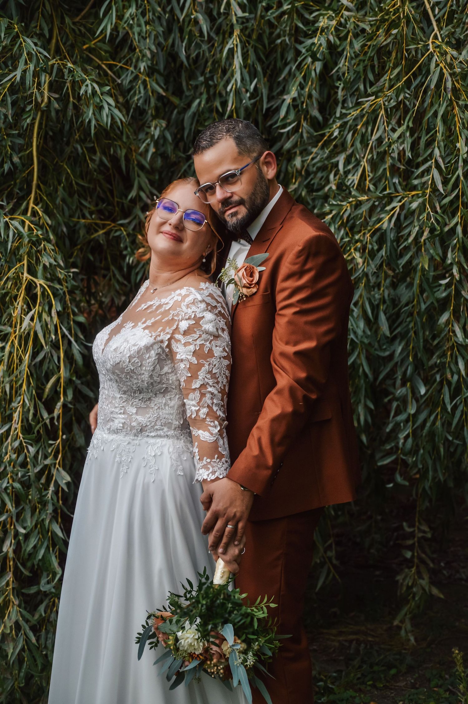 Wedding couple poses against willow tree backdrop in rust suit and white lace gown.